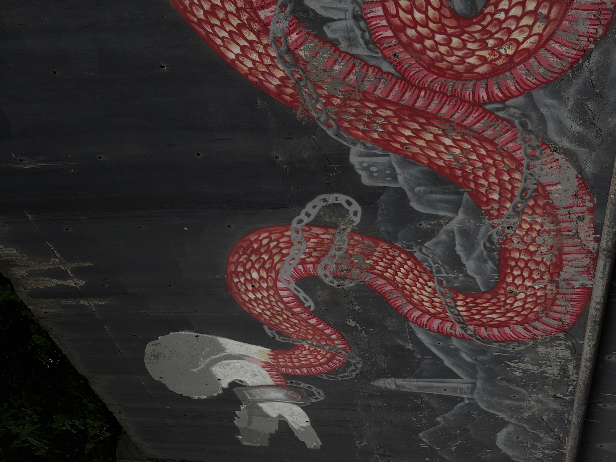 A red, scaled serpent is painted on a dark gray concrete wall. A silver-colored chain is wrapped around the snake in multiple places. The painting appears to be on the underside of a structure, possibly a bridge or overpass. Vegetation is visible at the lower left corner. The location is Tbilisi, Georgia.