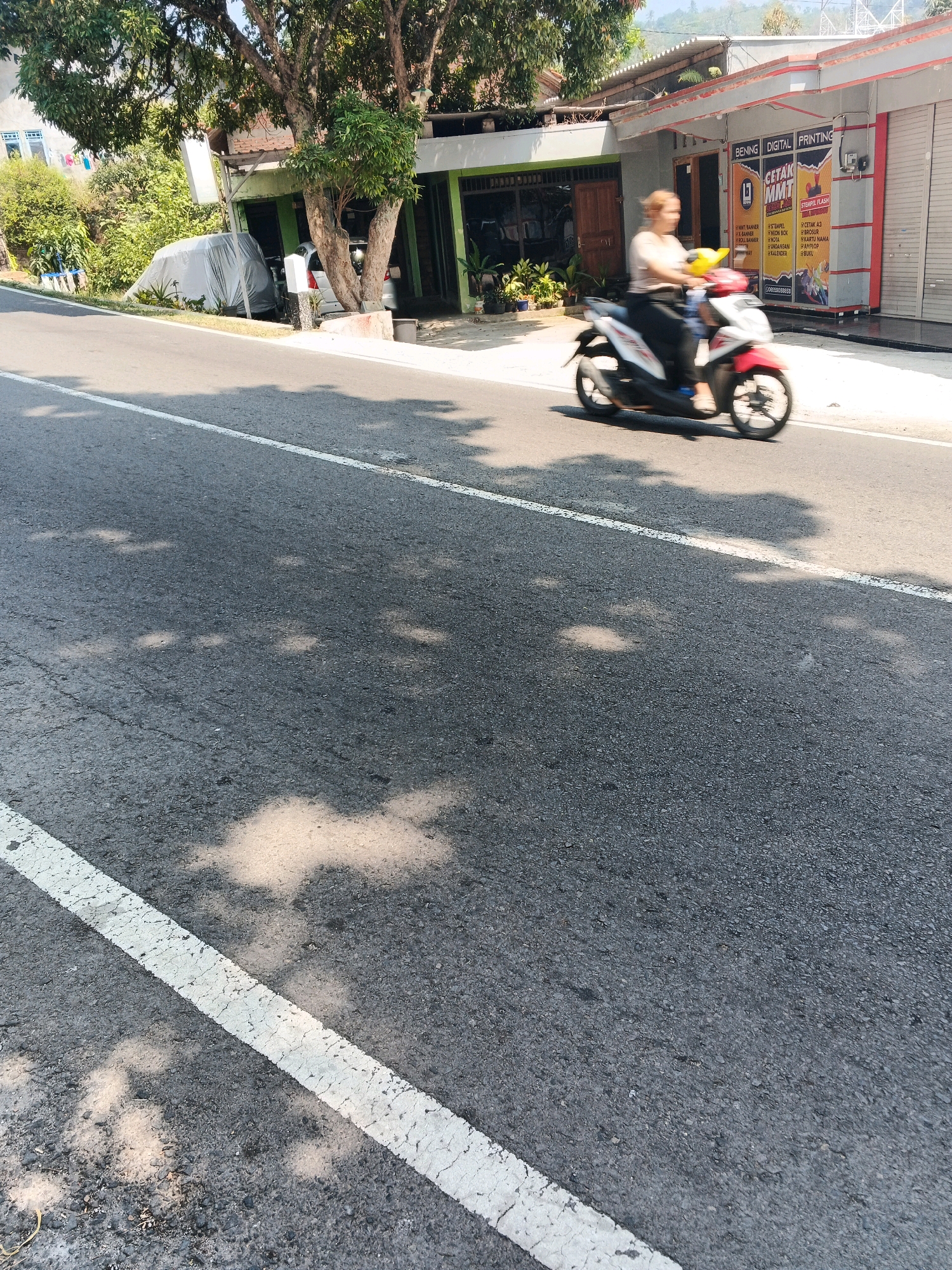 In Pakopen, Indonesia, a woman is riding a red and white scooter on a paved road. She is wearing a light-colored shirt and dark pants. The scooter has a yellow handlebar accessory. The road has a white line marking the lane. On the right side of the road, there is a building with a gray facade and red trim, featuring signage for "BENING DIGITAL PRINTING" and "CETAK MMT." Rolling metal shutters are visible on the building. Trees with lush green leaves are present to the left of the road, casting shadows on the asphalt. A gray tarp covers an object to the left of the road, partially obscuring a white car behind it. The background shows a hilly landscape with some buildings. The image is taken during the daytime, with bright sunlight creating distinct shadows.