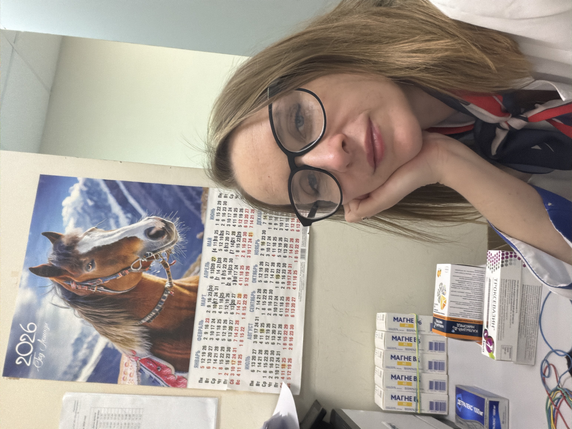 A woman with blond hair and glasses rests her head on her hand in front of a calendar for the year 2026 and a stack of medication boxes. The calendar has a picture of a horse and Cyrillic lettering indicating the year of the horse. The medication boxes are labeled in Cyrillic as well and include "MAGNE B6" and "DETRALEX 1000mg". The woman is wearing a white garment and a scarf. The background is a plain wall. There is no explicit information about the city of Zaprudnya.