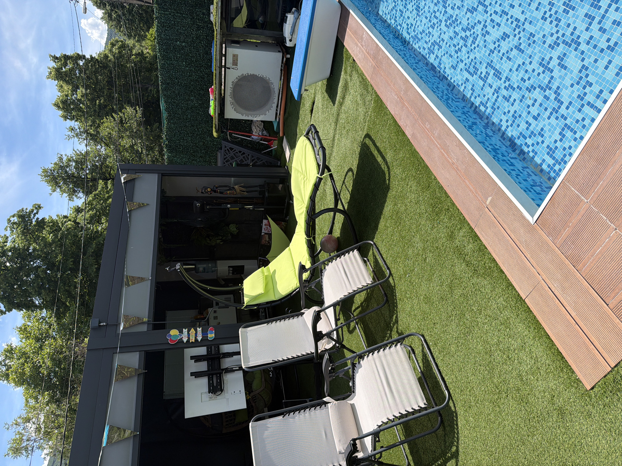 A bright, sunny day illuminates an outdoor recreational area in Sofia, Bulgaria. The scene features a swimming pool with blue mosaic tiles, bordered by a wooden deck. Adjacent to the pool is a patch of artificial turf, serving as a lounging area. Several lounge chairs are set up on the turf, with two light-colored chairs positioned in the foreground, and a vibrant green chaise lounge in the middle ground. Behind the lounge chairs, a modern, dark-colored structure with a flat roof is visible, adorned with triangular bunting flags. Inside the structure, a large white flat-screen television mounted on a stand is prominent. A hanging swing chair with a green cushion is suspended from the structure. Further back, a white air conditioning unit sits on a table, with various items on and around it. The background consists of lush green trees and a clear blue sky, suggesting a warm climate. The overall impression is one of a well-maintained private outdoor space designed for relaxation and entertainment.