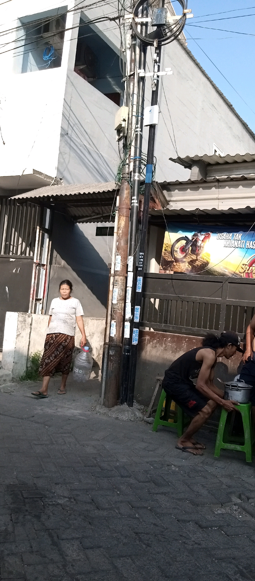In Wadungasri, Indonesia, a woman walks on a paved street carrying a large plastic water jug. She is wearing a white t-shirt and a patterned skirt. To her right, a man is seated on a green plastic stool, attending to a pot on another stool. He has dark, curly hair and is wearing a black sleeveless shirt and shorts. Behind them, a utility pole stands adorned with various cables and small electronic boxes. A building with a corrugated metal roof is visible behind the woman, and a banner with an image of a motorcycle and Indonesian text hangs on a fence. The scene takes place under a clear blue sky with bright sunlight casting shadows.