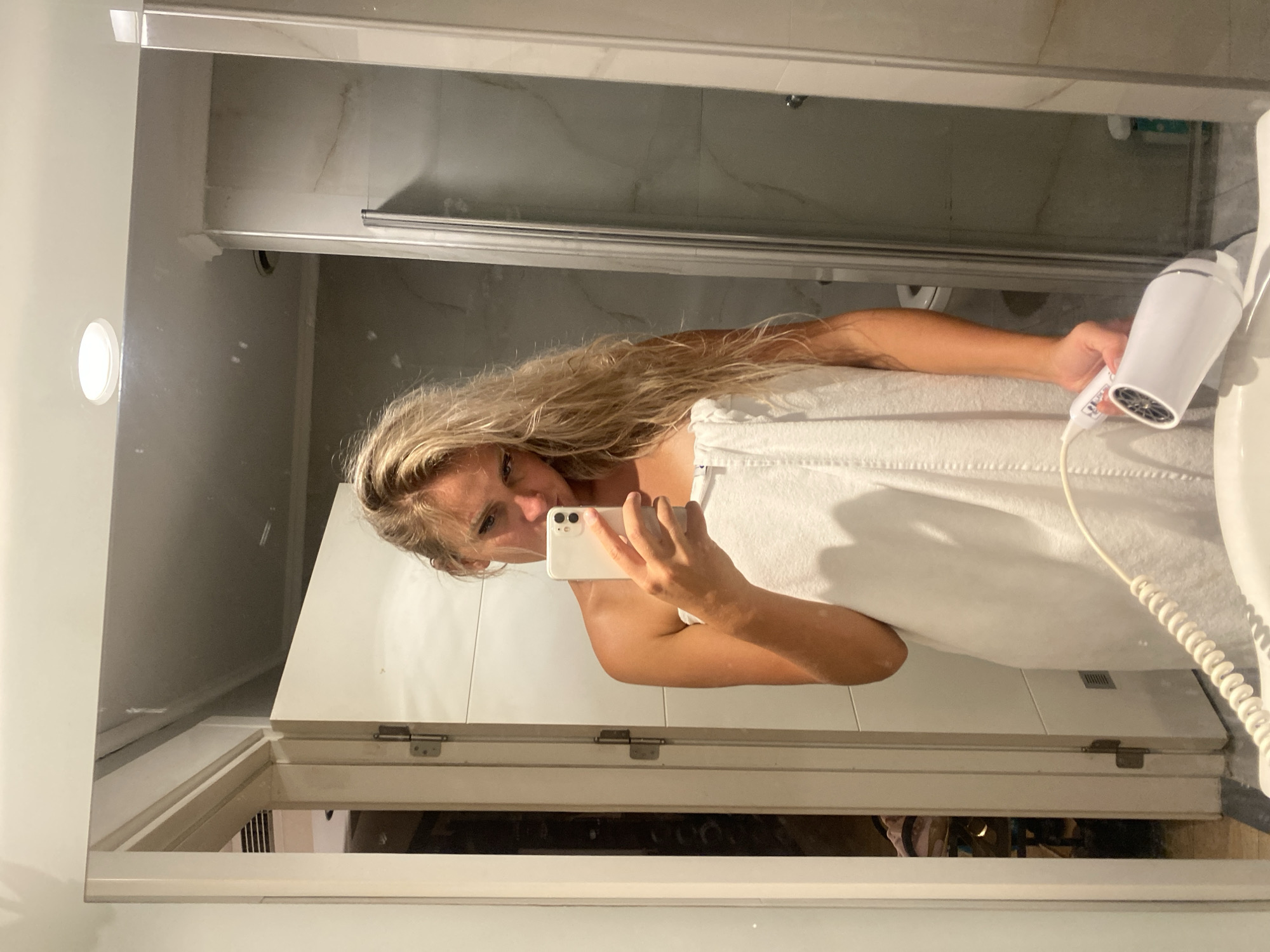A woman, appearing to be in her early 20s, stands in front of a mirror, taking a selfie with her phone. She is wearing a white towel wrapped around her body, covering her from the chest down. Her blonde hair is long and slightly damp, reaching down her back. She is holding a white hairdryer in her right hand. The background suggests a bathroom environment, with a shower stall visible to her right and a door to her left. The overall impression is that of a post-shower scene.
