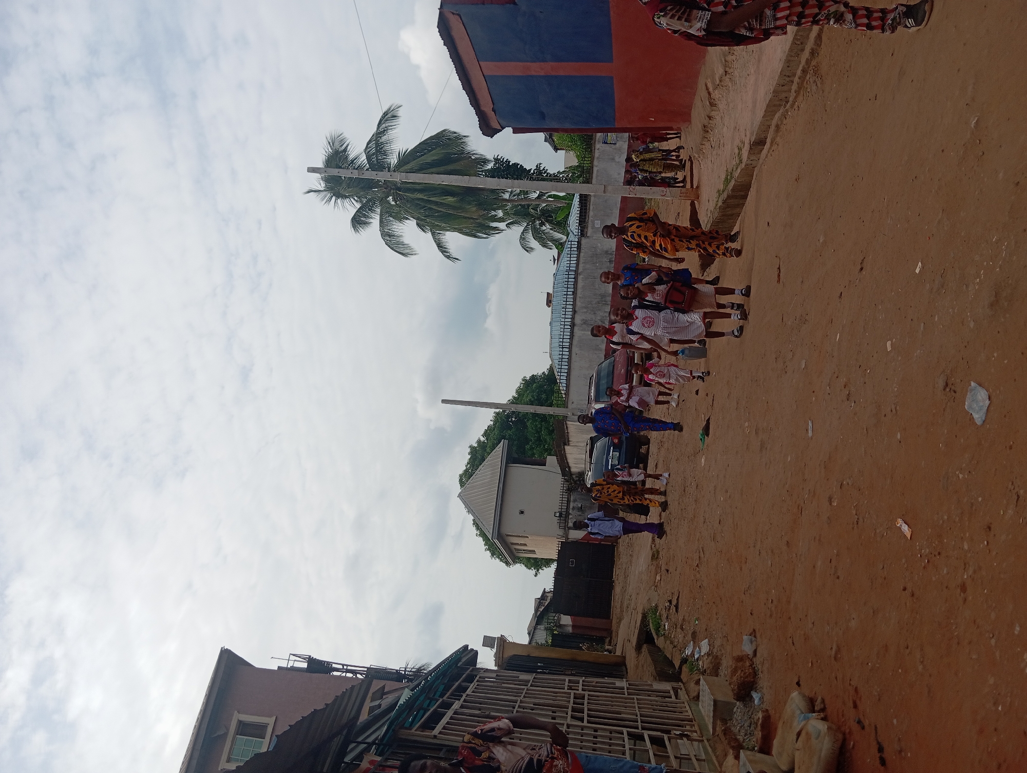 The image depicts a street scene in Nkpor, Nigeria. The street is unpaved, with a reddish-brown surface. Buildings are visible on both sides of the street, constructed with varying materials and colors. A palm tree stands tall in the background. A group of people, including adults and children, are walking in a line along the street. The individuals are wearing a mix of traditional and casual clothing. A vehicle is parked next to the people. The sky is overcast with clouds. Debris and construction materials are scattered along the street.