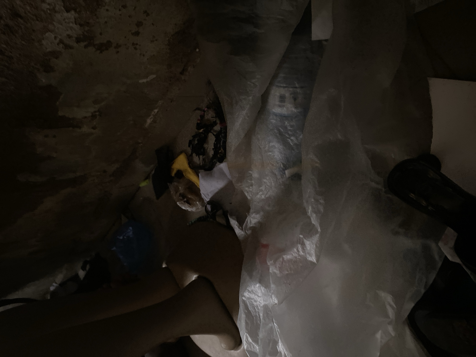 The image depicts a dimly lit, cluttered interior space. On the left, a mannequin's leg and foot are visible, partially covered by a large sheet of crumpled white plastic. Scattered amidst the plastic are various discarded items including a blue plastic bag, a yellow object, and dark-colored clothing with intricate patterns. The background appears to be a rough, textured wall, possibly made of concrete or mud, with some lighter colored patches. The overall impression is one of disarray and abandonment. The location in Jauro Yinu, Nigeria, suggests an urban or peri-urban setting, though the specific context of this interior space remains unclear without further information.