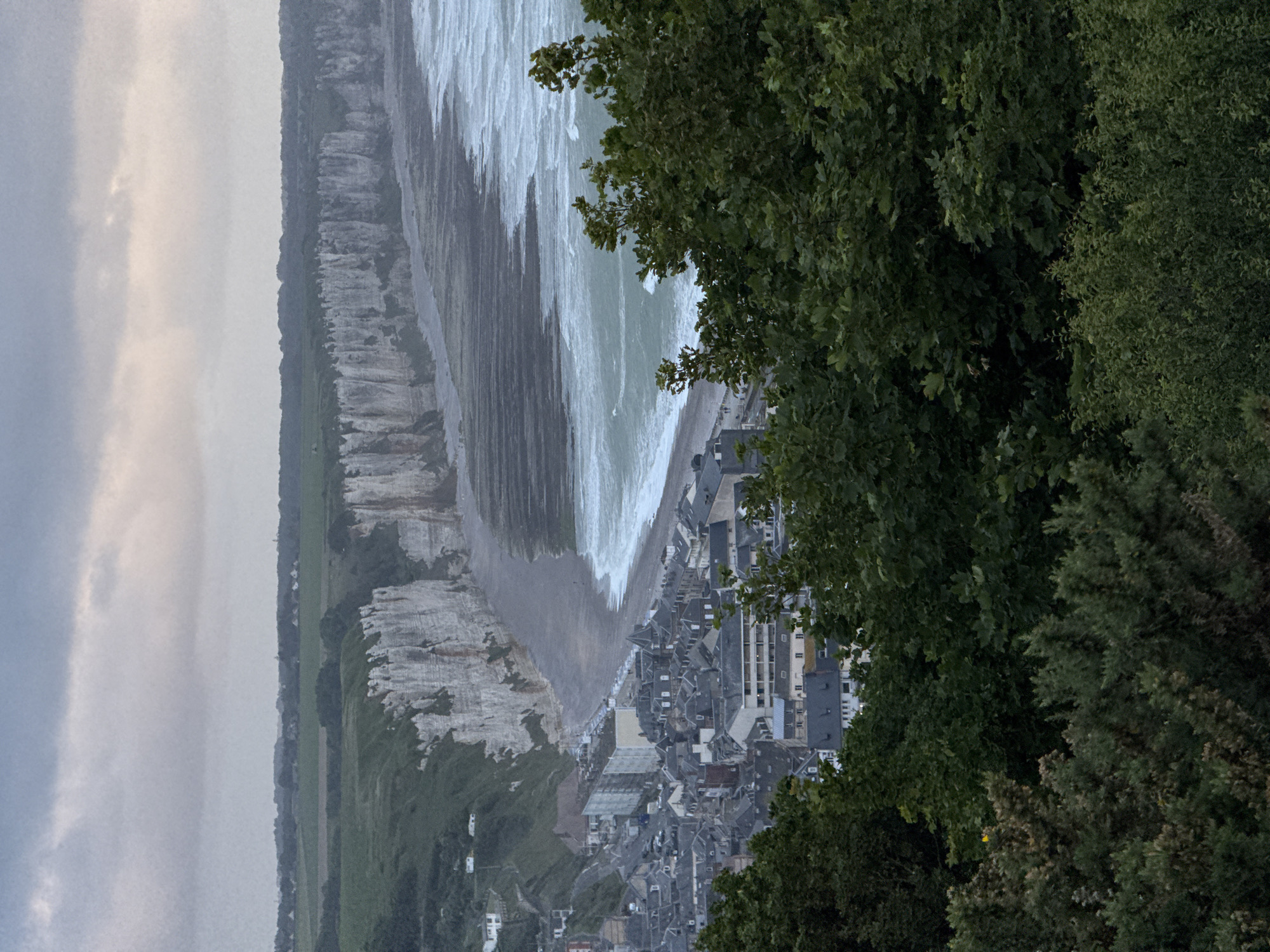 The photograph, originally rotated 90 degrees clockwise, captures an expansive, elevated view of the coastal town of Fécamp, France, under an overcast sky.
The scene is dominated by a dramatic landscape where towering white chalk cliffs stretch horizontally across the left side, meeting a dark, stony beach. Beyond the cliffs, a rural landscape with green fields and scattered distant structures extends under a pale grey, cloudy sky.
The greenish-grey sea occupies the central and right portions of the image, with prominent white waves actively crashing onto the shore, indicating an energetic tide or windy weather.
Nestled directly behind the beach, the town of Fécamp unfolds with a dense arrangement of multi-story buildings. These structures, characterized by their dark roofs (likely slate or tile) and traditional European coastal architecture, line the coastline. Due to the distance, no specific human activities or individual people are discernible, but the tight cluster of buildings clearly points to a well-established community.
Lush green trees and dense foliage frame the immediate foreground on the right, partially obscuring parts of the town and the sea. This vegetation suggests the viewpoint is from a high, wooded area overlooking the coast. The soft, diffused lighting from the heavy cloud cover gives the scene a muted yet powerful ambiance, characteristic of a cloudy day on the Normandy coast. There is no visible text in the image.