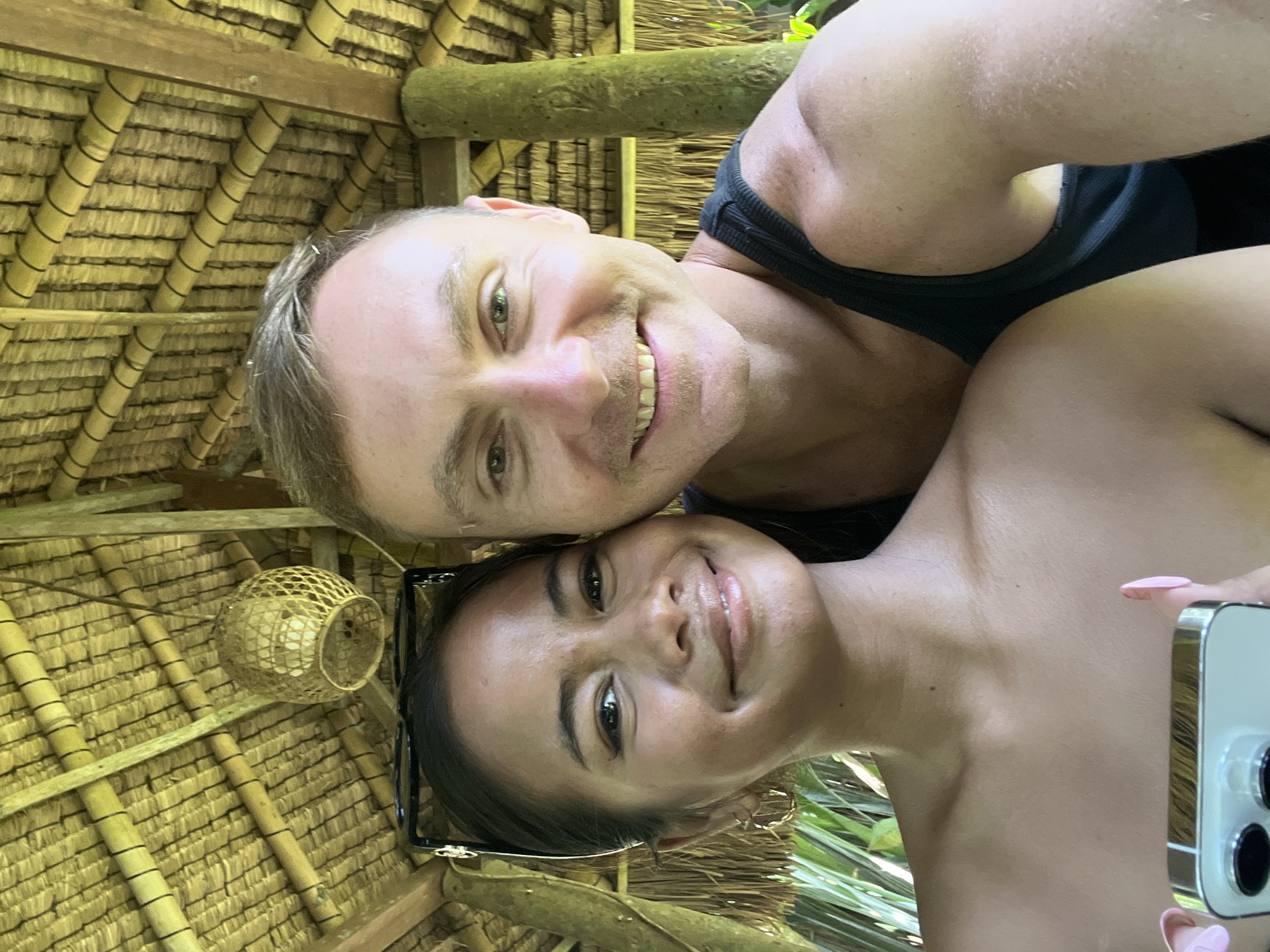 A close-up, selfie-style photo shows a couple smiling at the camera, positioned under a thatched roof structure. The woman on the left has dark hair pulled back and wears hoop earrings. The man on the right has short, light brown hair and is wearing a black tank top. They are leaning into each other, suggesting a close relationship. The background reveals a rustic, tropical environment with bamboo beams and palm leaves visible. A woven light fixture hangs from the ceiling. The scene suggests they are outdoors in a warm climate, possibly a resort or a natural dwelling. The photo was taken in Blahbatuh, Indonesia.