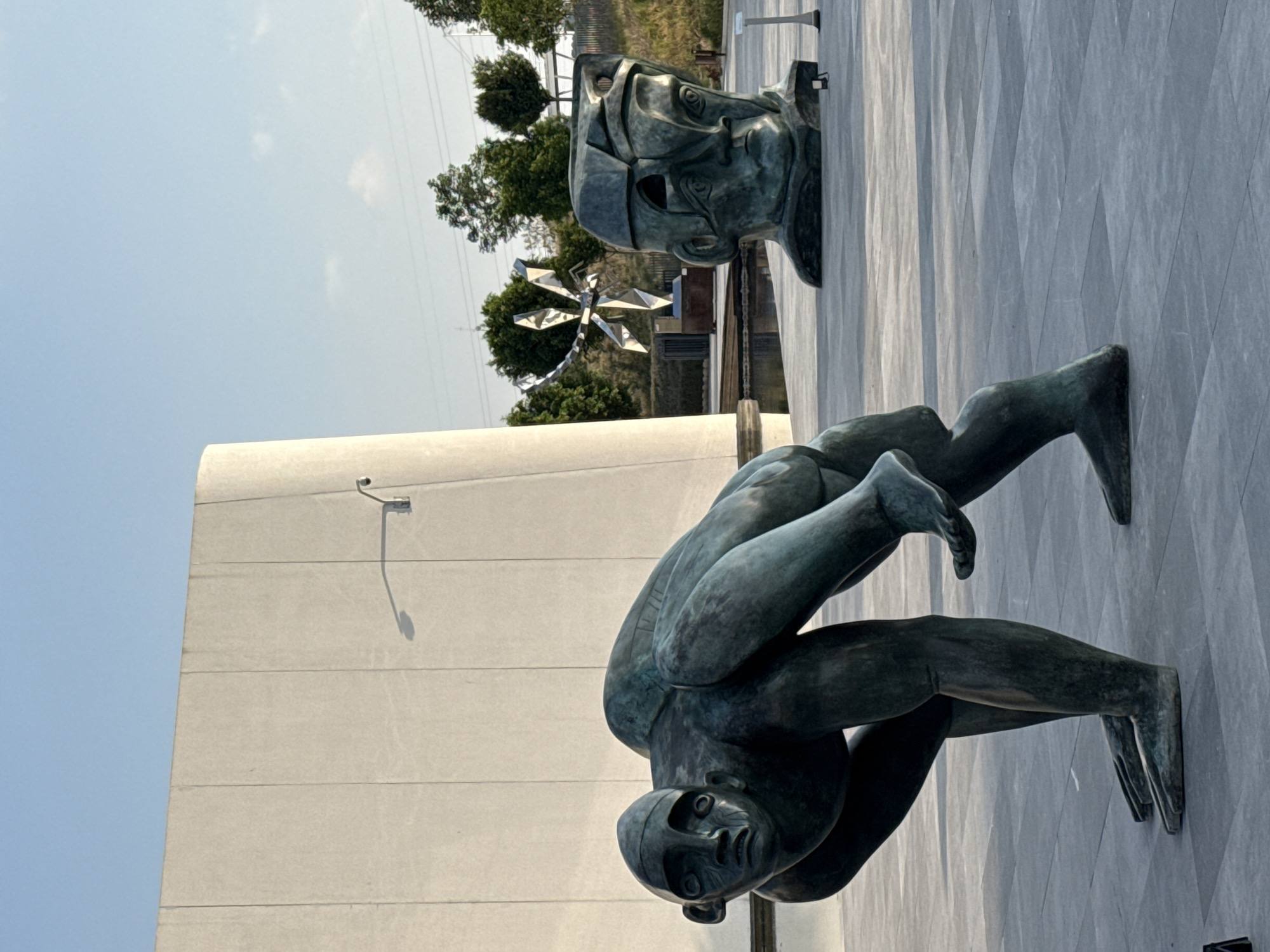 The image shows two bronze sculptures mounted on exterior walls in San Bernardino Tlaxcalancingo, Mexico. One sculpture depicts a person bent over with their legs extended up to another sculpture above. The second sculpture is a stylized head. Both sculptures appear to be attached to modern, light-colored walls. The background includes trees, an abstract metal sculpture resembling a dragonfly, utility poles, and a clear sky. The surfaces around the sculptures are paved. The image is taken from a low angle, exaggerating the height of the walls and sculptures. A security camera is visible on the white wall behind the contorted body.