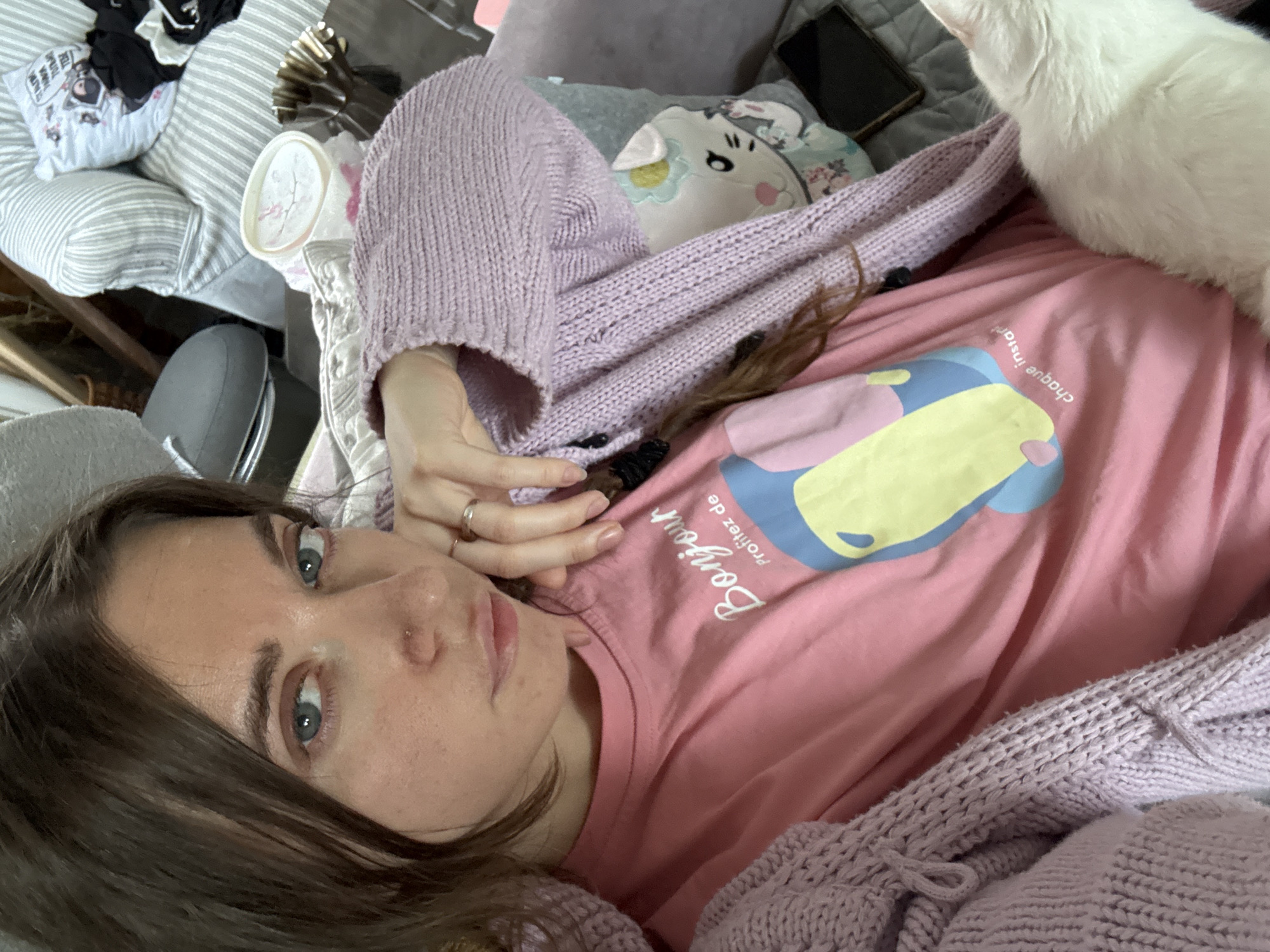 A woman with brown hair and blue eyes is taking a selfie in a room. She is wearing a pink t-shirt with a graphic and French text, and a purple knitted cardigan. She is resting her chin on her hand, which is adorned with two rings. A white cat is visible in the lower right corner of the image. In the background, a striped sofa and a pillow with a cat illustration are present. The environment appears to be a living room.