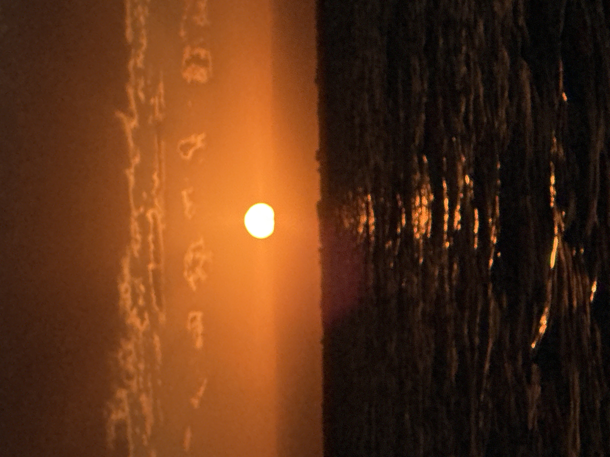 The image shows a sunset over a body of water. The sun, a bright orange orb, is low on the horizon, casting a warm glow across the sky and reflecting on the water. The sky is filled with wispy clouds, tinged with shades of orange and brown. The water is dark and choppy, with streaks of light from the setting sun shimmering on its surface. The scene evokes a sense of tranquility and natural beauty, typical of a sunset over a lake or ocean. There is no indication of the city of Kokshetau, Kazakhstan, in this image.