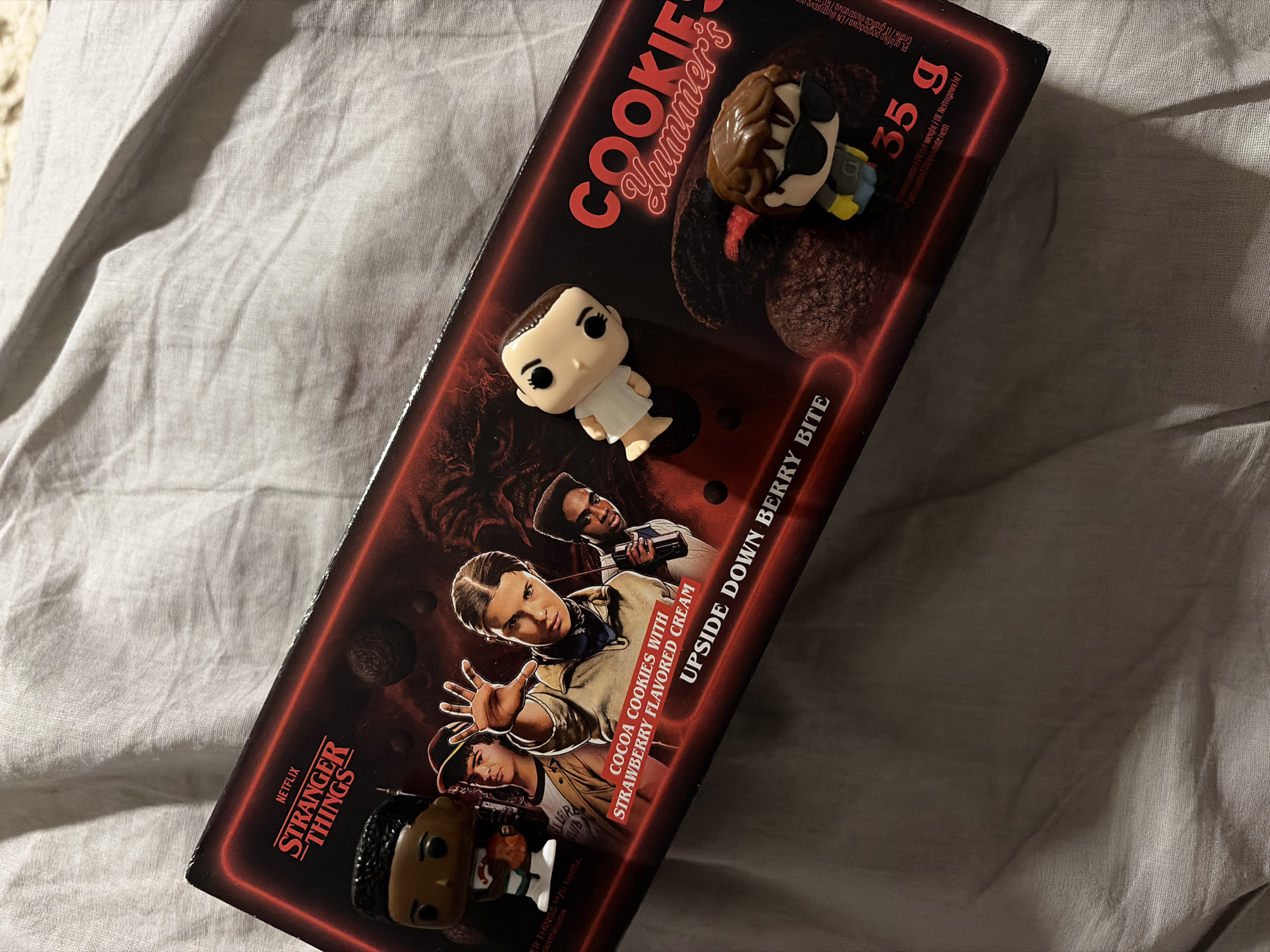 The image displays a collection of Stranger Things-themed merchandise, including a cookie package and three Funko Pop-style figurines, resting on a light-colored, wrinkled fabric surface. The scene takes place indoors, likely on a bed or table, in Perm, Russia, under dim lighting that creates soft shadows.
The prominent object is a rectangular cookie package from "COOKIES Yummer's," adorned with the Netflix "STRANGER THINGS" logo and character art featuring Eleven, Lucas, and Dustin. The product is identified as "COCOA COOKIES WITH STRAWBERRY FLAVORED CREAM" and marketed as an "UPSIDE DOWN BERRY BITE." The package indicates a weight of "35 g" and features a dark background with striking red outlines and text. Visuals of the dark cocoa cookies are also present on the packaging.
Three Funko Pop-style figurines are strategically placed around the cookie box. One figurine, resembling Eleven in her white dress, is positioned on the top left of the package. Another, partially visible on the top right, appears to be a monstrous character or a highly detailed character with a complex headpiece. A third figurine, depicting Lucas Sinclair, is located at the bottom left, also partially out of frame.
The arrangement is static, showcasing themed collectibles and snack food, appealing to fans of the series. The fabric provides a soft, textured background, contrasting with the glossy packaging.