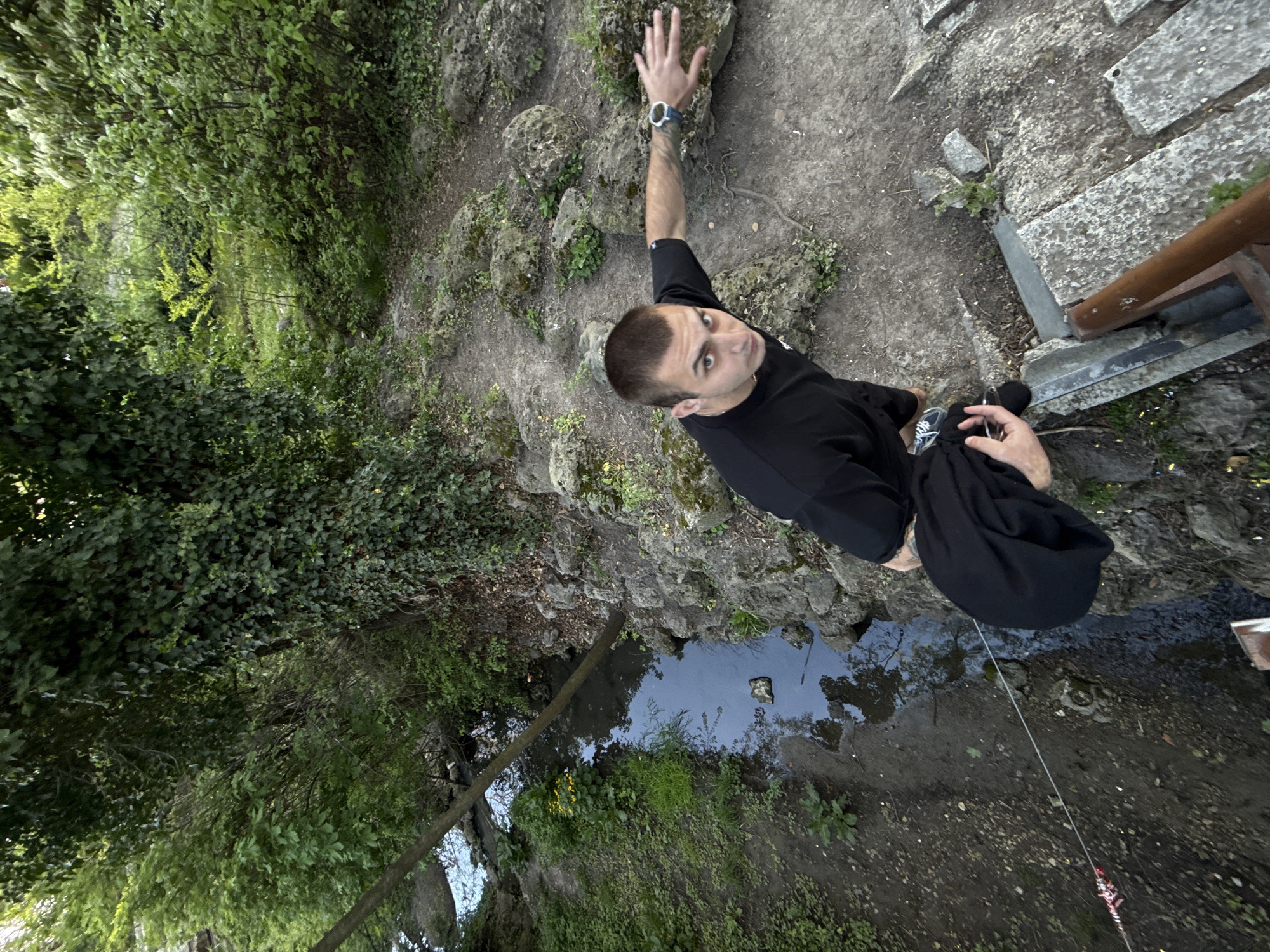 A medium, wide-angle shot looking downward at a man in an outdoor, natural environment. The man is looking up at the camera. He has dark, cropped hair, and is wearing a black t-shirt, black pants, and athletic shoes. He is standing beside a small pond or stream with his right arm extended, palm open, touching a cluster of mossy rocks and ground. A watch is visible on his wrist. To his left is a steep embankment covered with green foliage. Behind him is a stone walkway or path, with sections of metal railing visible on the right side of the frame. In the pond are some fallen limbs and rocks, and the pond reflects the sky. A thin, possibly fishing line, is visible going from the man to the ground toward the bottom right of the frame.
The scene appears to be taken in Varna, Bulgaria, near the Asparuhovo Bridge. The vegetation type and rock composition are consistent with that region. The structure of the path and small bodies of water are indicative of the park or gardens nearby.