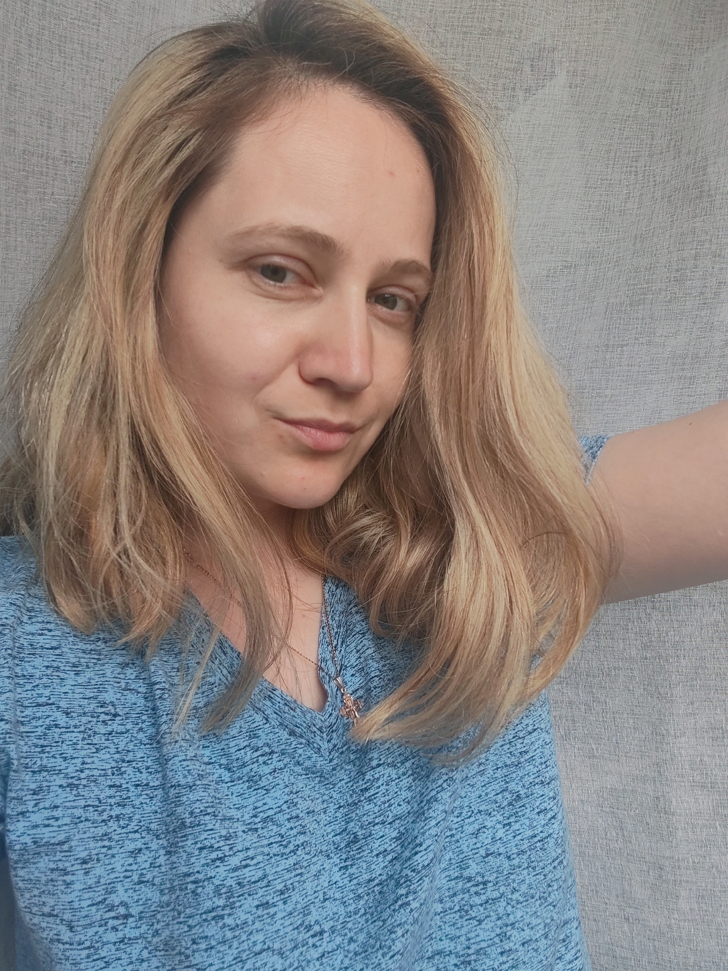 The image shows a close-up selfie of a woman with blonde hair, wearing a blue t-shirt. She is looking towards the camera with a slight smile. The background is a neutral, light-colored fabric. The lighting appears to be soft, suggesting it could be daytime. The woman is wearing a necklace with a small pendant. No text is visible in the image. The context provided indicates the location is Sokol, Russia.