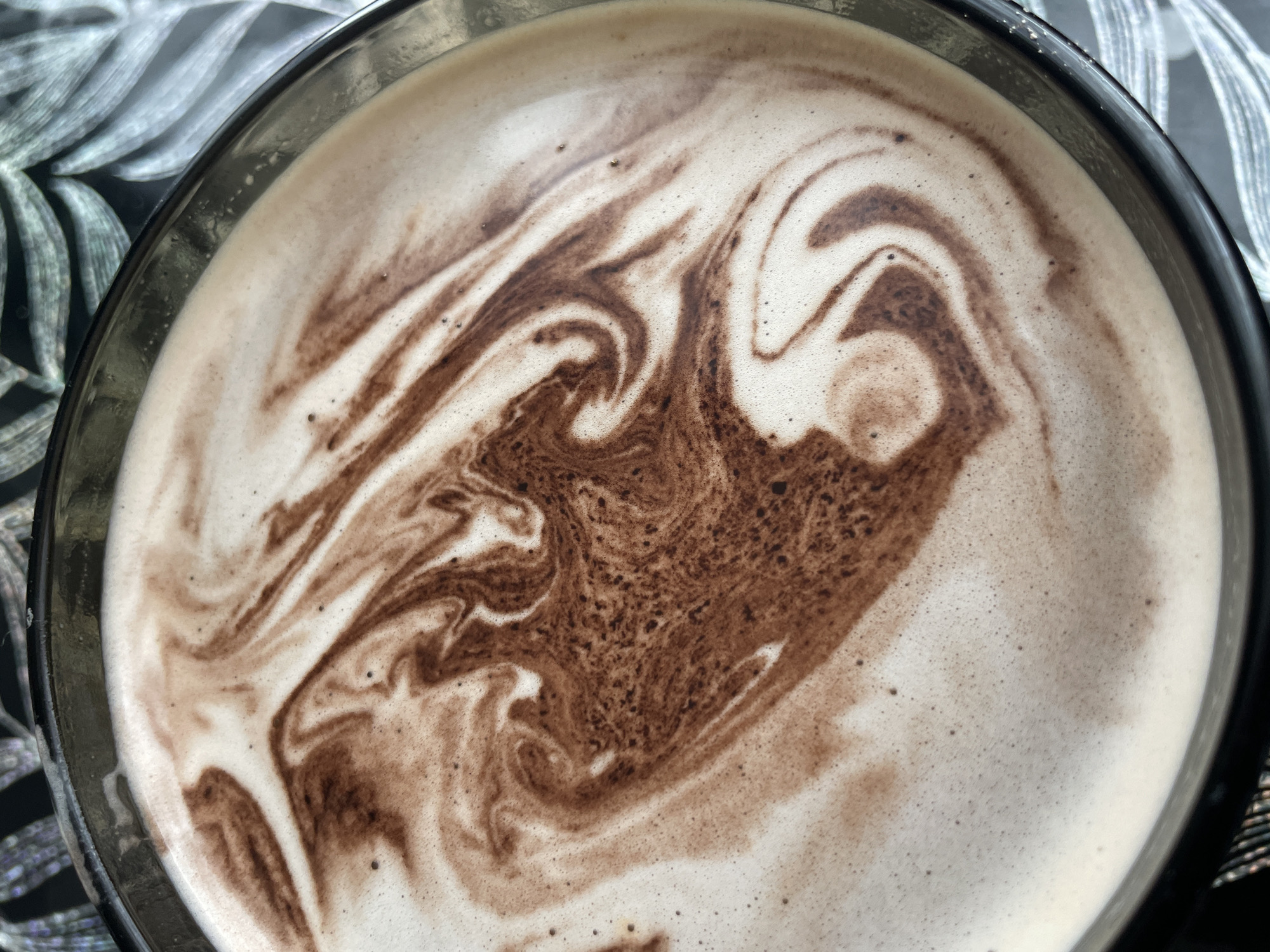 A top-down view of a glass cup filled with a light-colored beverage featuring swirls of dark brown chocolate. The cup is set on a surface with a patterned cloth underneath. The pattern on the cloth appears to be abstract leaf shapes in shades of gray.
There is no information available about the location of the image being in Chaikovsky, Russia, or any people, subjects, objects, actions, or interactions within the image beyond the cup of beverage.