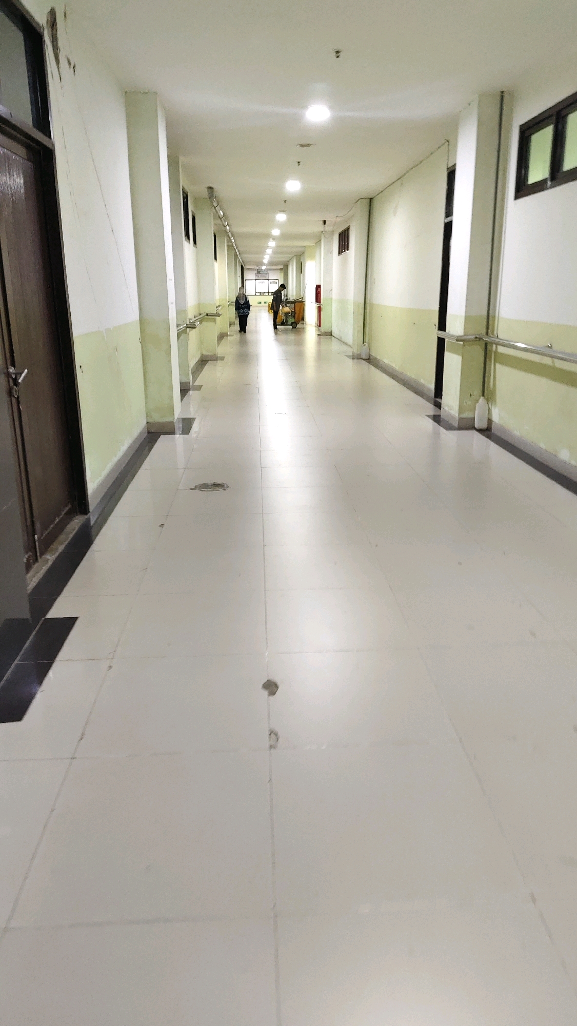 The image shows a long, brightly lit hospital corridor in Banjarmasin, Indonesia. The corridor has a white tiled floor and pale green and white walls. Fluorescent lights are evenly spaced along the ceiling. On the left side of the corridor, there is a dark wooden door and a railing. On the right side, there are windows and another railing. Two individuals are visible in the distance. One, wearing a headscarf, walks away from the camera. Another person, possibly a hospital worker, is pushing a yellow cart. The scene is clean and orderly, typical of a healthcare facility.