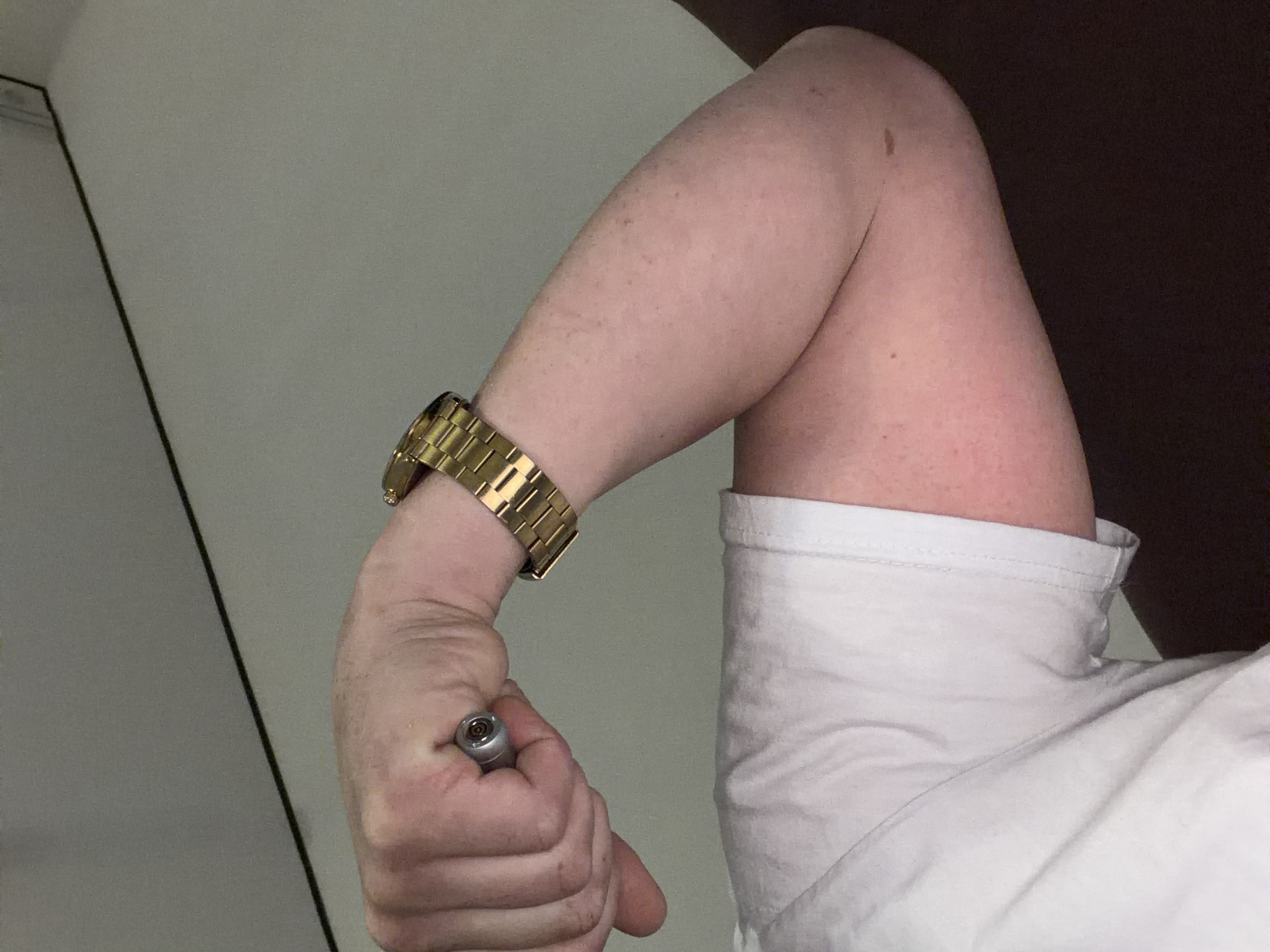 A close-up shot depicts a person's flexed arm, bent at the elbow, with a gold watch on their wrist. The person is holding a silver object, possibly a vape pen or similar cylindrical device, in their closed fist. The subject is wearing a white t-shirt. The background is a neutral wall with a dark section on the right side of the image. There is no indication of the location being Chaikovsky, Russia.
