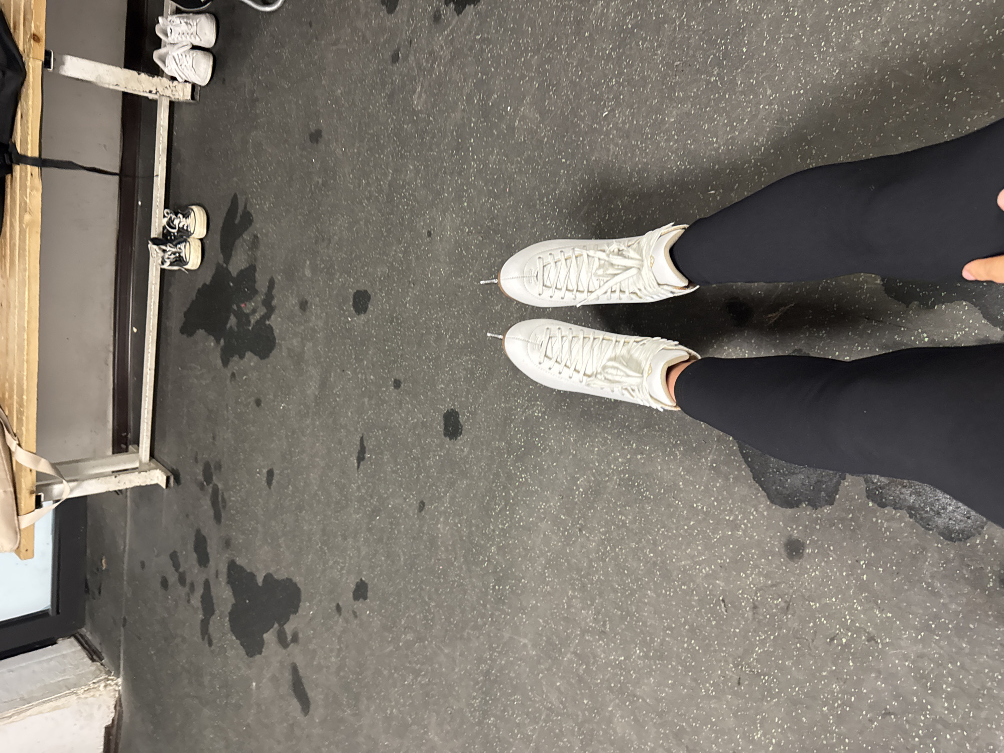 A high-angle shot shows a person wearing white figure skates and black leggings standing on a gray floor with several dark, irregular patches, possibly water or stains. To the left is a locker room with a bench, where pairs of shoes are visible. The scene appears to be inside an ice rink in the city of Unknown, Unknown.