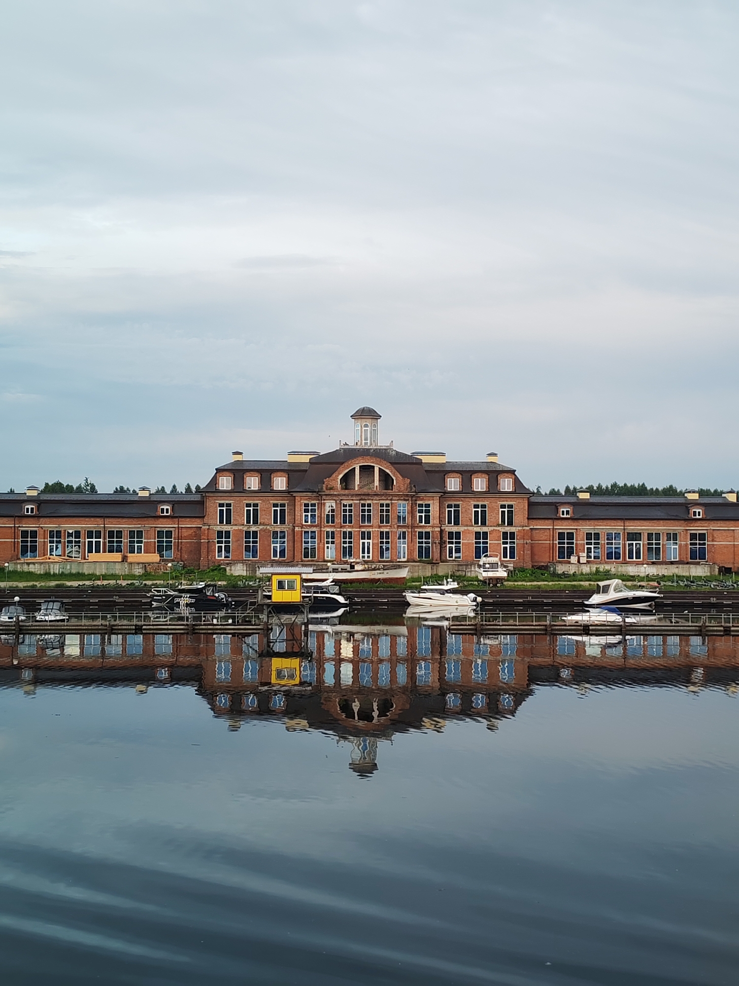 The image depicts a grand, multi-story brick building with a dark roof and numerous large windows, situated on the waterfront. The building is symmetrical in design, featuring a central section with an arched facade and a smaller domed structure on top. A reflection of the building and the boats docked in front of it is visible in the calm water of the river or canal. Several boats, including speedboats and what appears to be a yellow lifeguard station or small building, are docked along a pier in front of the main structure. The surrounding area is lined with trees, and the sky above is overcast. The scene appears to be a modern development or a refurbished historical industrial site. The text indicates the location is Gorodnya, Russia. There are no people visible in the image.