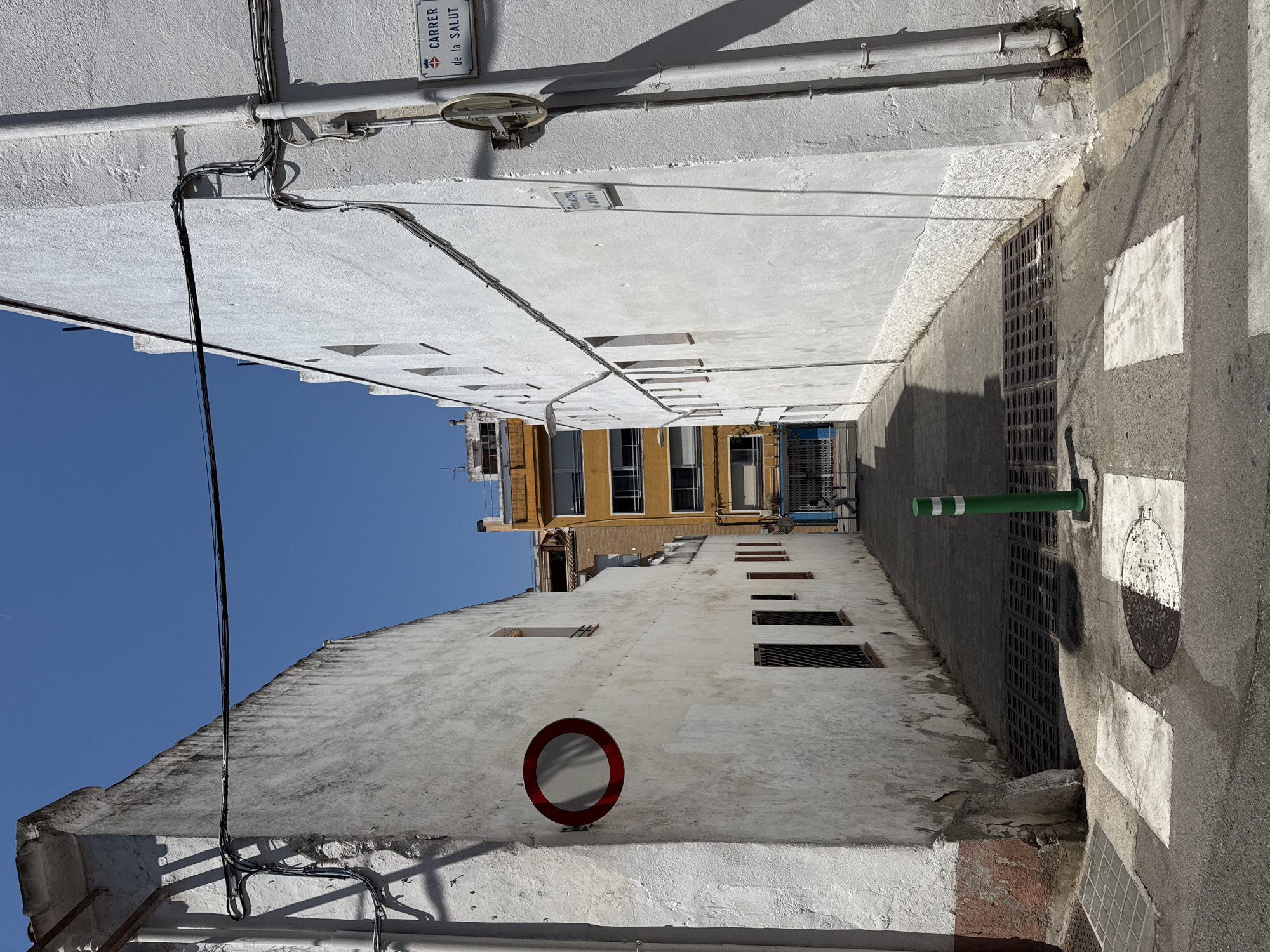 The image shows a narrow, sun-drenched alleyway in Blanes, Spain, with tall, white-washed buildings on either side. The alleyway has a paved surface with a crosswalk painted on it. A green bollard with white stripes stands near the crosswalk. On the left building, a circular red and white traffic sign is mounted on the wall. On the right building, a street sign reads "CARRER de la SALUT". Electrical wires are strung across the alley and run along the sides of the buildings. In the background, a yellow multi-story building is visible, along with glimpses of other buildings and a clear blue sky. There is one person walking in the distance at the end of the alley.