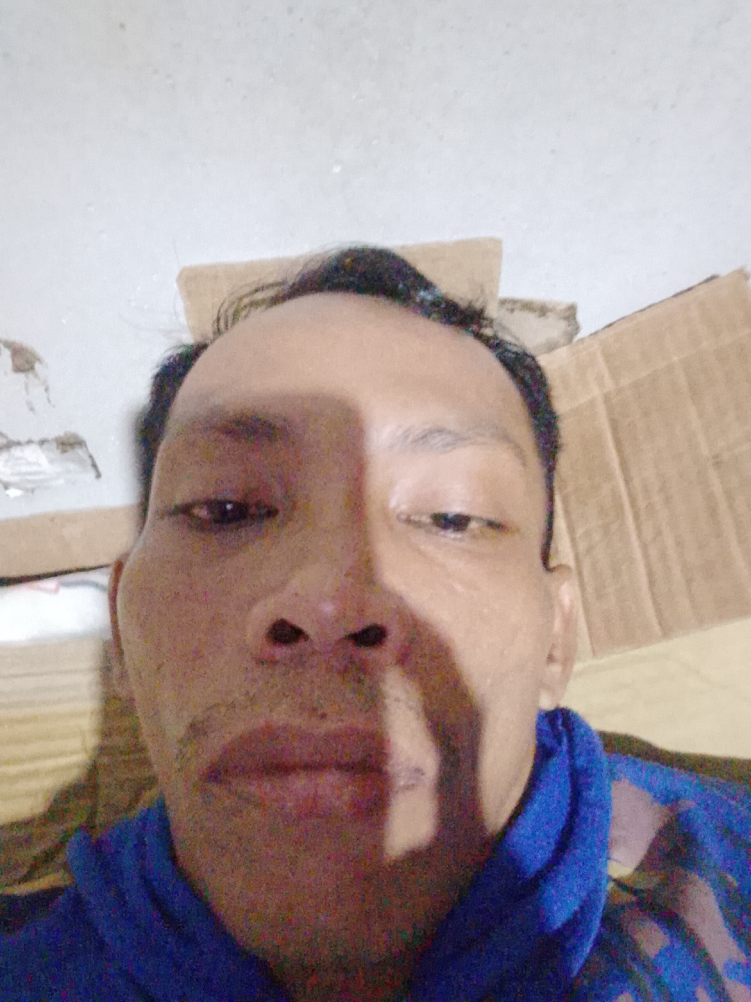 A close-up, upward-angle selfie of an adult male with dark hair, brown eyes, and tan skin is presented against a cluttered background. The man is wearing a blue top with a collar. Behind the man, portions of cardboard boxes and what appears to be a white wall with some damage or discoloration are visible. The lighting is uneven, creating strong shadows on the man's face. The picture potentially was taken in Bukit Ketok, Indonesia.
