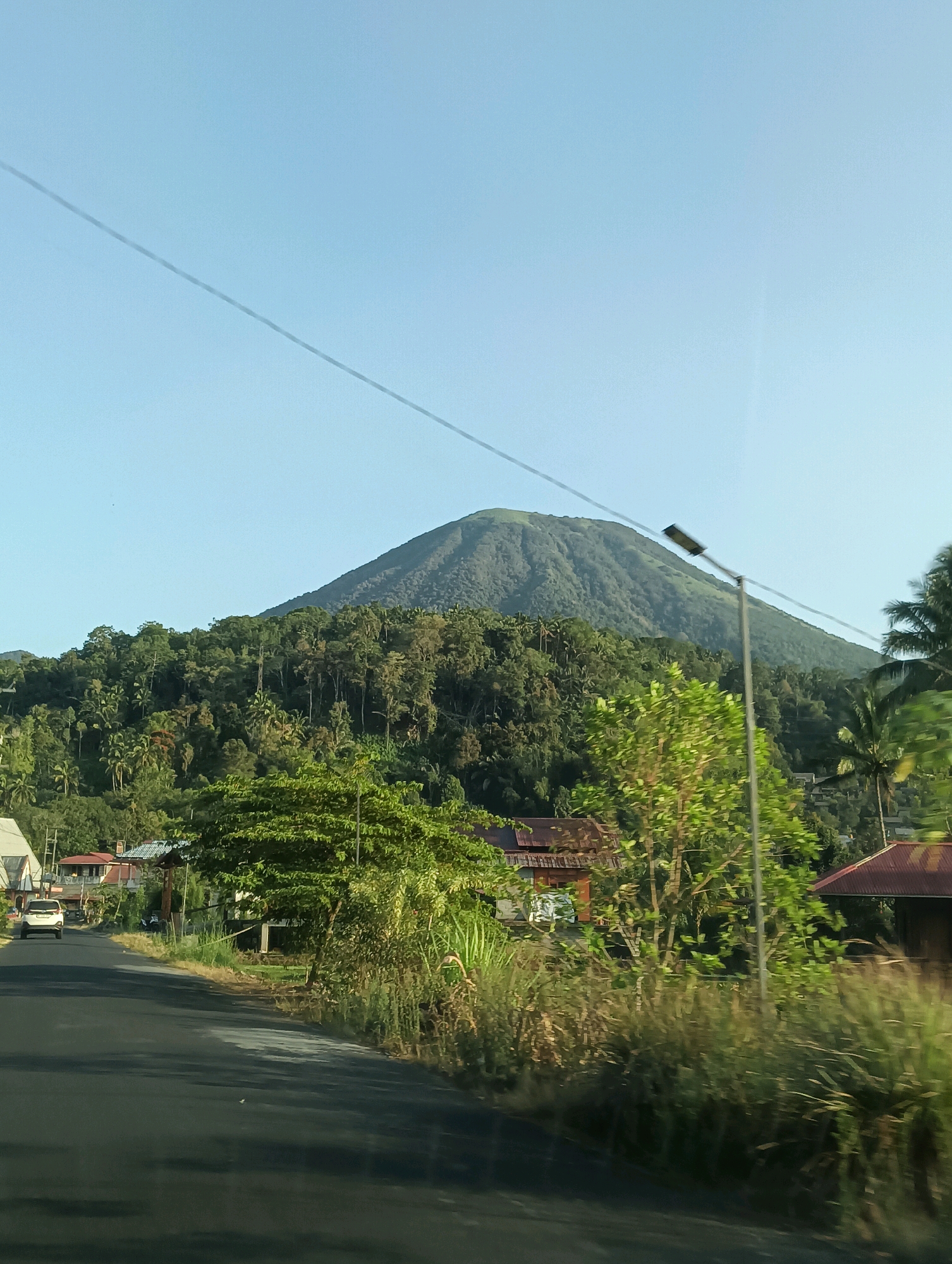 The image captures a scenic view of a mountain, likely a volcano, dominating the background. The mountain is covered in lush green vegetation, indicative of a tropical environment. In the foreground, a road curves gently, with a white vehicle visible in the distance on the left side. Along the roadside, there are houses with red tiled roofs, interspersed with various trees and tall grass. A lamppost stands tall on the right side of the road, with power lines running diagonally across the clear blue sky. The overall impression is of a rural or semi-rural setting, possibly in or around Tomohon, Indonesia, known for its volcanic landscape and natural beauty. The lighting suggests it is daytime, with the sun casting shadows on the road. There are no people clearly visible in the image.