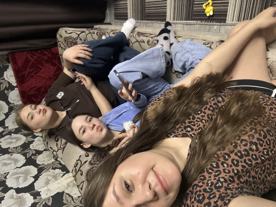 Three young females are seated on a patterned sofa in an indoor residential setting in Ольховка, Russia. The foreground shows a female with long brown hair, wearing a leopard-print top, holding a smartphone to take a selfie. She has a slight smile. Adjacent to her, a second female with braided hair, wearing a light blue zip-up hoodie and light blue jeans, poses with a "peace" sign and a duck face, holding a smartphone in her right hand. She wears black and white spotted animal-themed slippers on her feet. In the background, a third female with light brown hair, wearing a dark brown polo shirt and dark blue jeans, looks towards the camera with a pouting expression, holding a smartphone in her right hand. She wears rings on her left hand and a white sock on her left foot. The sofa has a light brown floral pattern. Behind them, a wall is covered with dark wallpaper featuring large white floral designs. A dark red textured pillow and a dark curtain are visible to the right. A patterned rug or carpet with a small yellow toy is visible on the floor to the right.