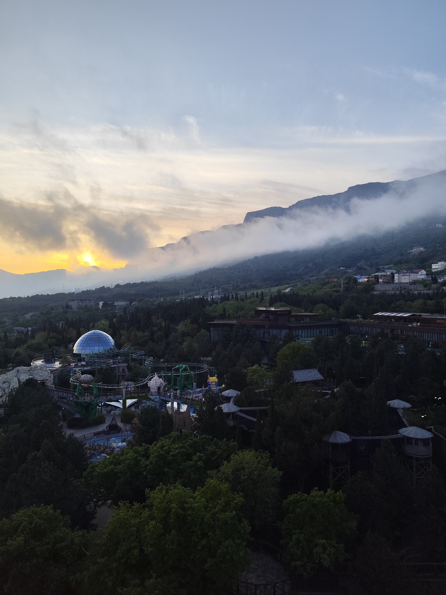 This image shows an elevated view of a resort area in Симеизский поселковый совет, Ukraine. The scene includes a mountain range partially obscured by fog. In the valley, there are structures including a large dome-shaped building, a roller coaster, hotel buildings, and forest. The sky is cloudy with the sun near the horizon.