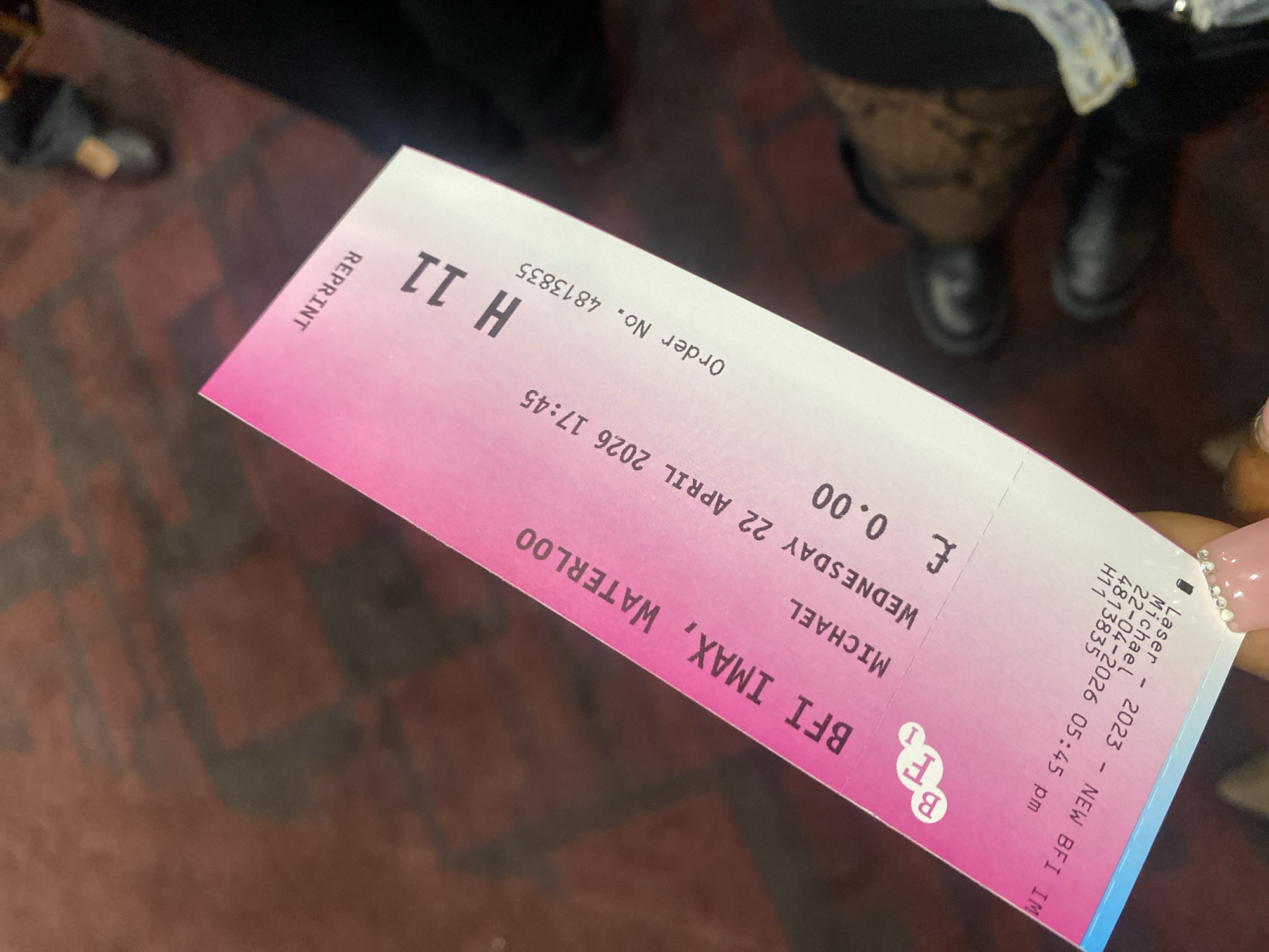 A close-up shot captures a pink and white cinema ticket held by a hand with neatly manicured, light-colored nails adorned with small rhinestones. The ticket is for "BFI IMAX, WATERLOO" and is dated "WEDNESDAY 22 APRIL 2026 17:45" (5:45 PM). It indicates the ticket holder as "MICHAEL" and the seat number as "H 11". A notable detail is the price, shown as "£ 0.00", and the word "REPRINT" clearly visible on the ticket. The order number "4813835" is also printed. In the blurred background, several people's legs and feet are visible, standing on a dark reddish-brown patterned floor, suggesting an indoor public space such as a cinema lobby or waiting area in London, United Kingdom. The image implies an activity of attending a film screening at the BFI IMAX.