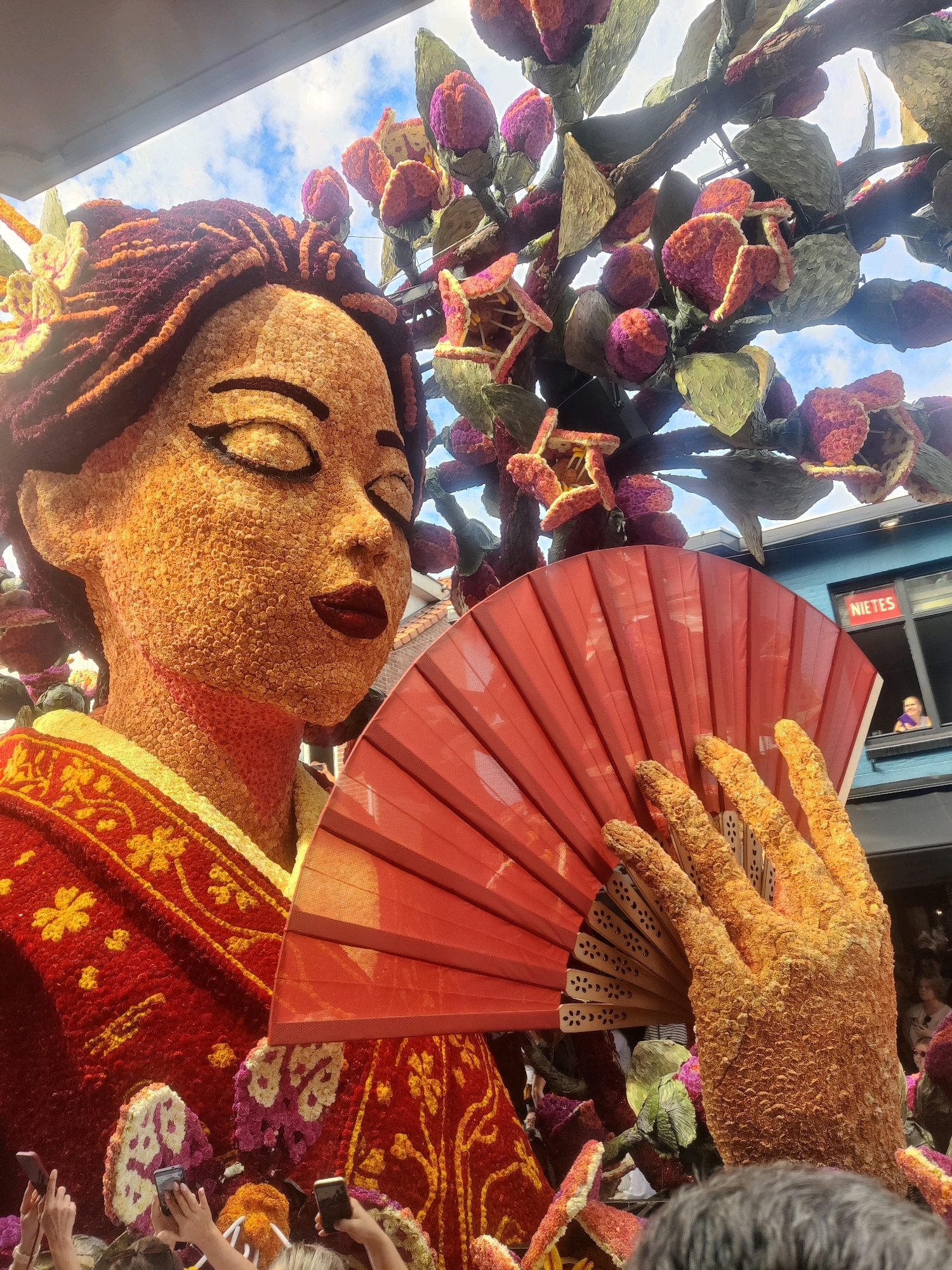 This is a medium-wide, eye-level, outdoor shot of a large floral float depicting a woman in traditional Japanese attire, presented in the city of Zundert, Netherlands. The float is adorned with an abundance of orange, yellow, red, and purple flowers, forming the woman's face, braided hair, hand, and elaborate kimono. Her eyes are closed, and she holds a bright red fan in her right hand, which is also made of flowers. The background features a blue building with a window displaying the word "NIETES" and a glimpse of a person inside. Below the float, the hands of several onlookers holding smartphones are visible, capturing the spectacle. The float is situated amidst other floral displays and greenery, set against a partly cloudy blue sky.