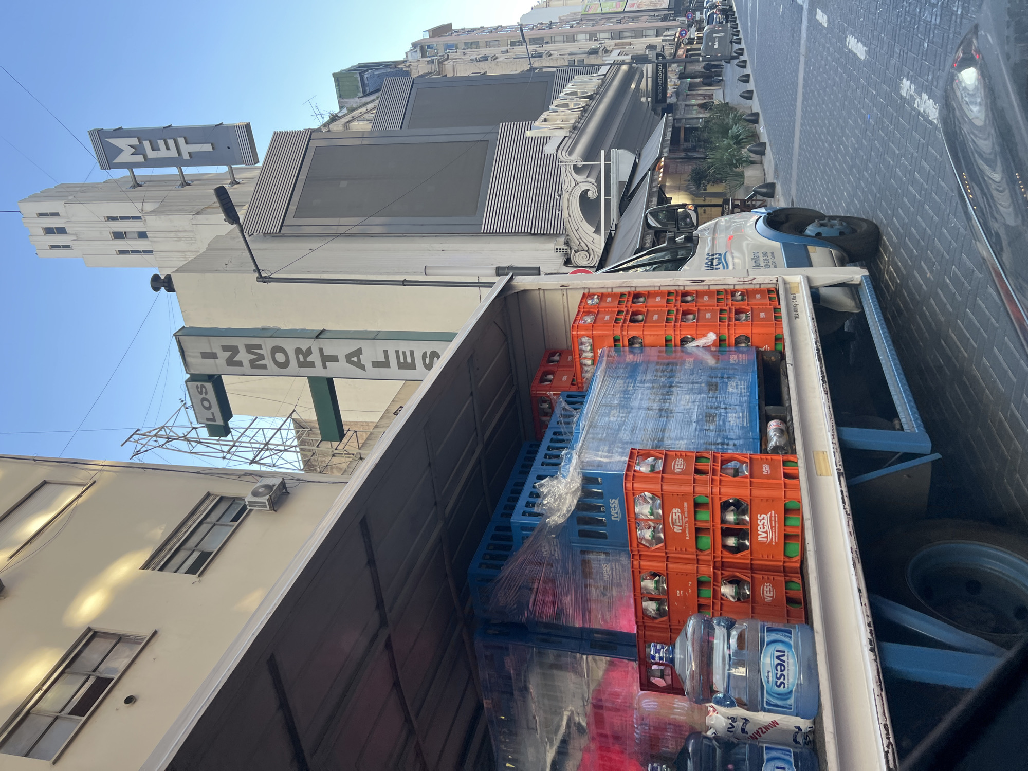 The image captures a street scene in Buenos Aires, Argentina, specifically near the Teatro Metrópolis (Metropolitan Theater). The view is from inside a delivery truck filled with orange and blue plastic crates of IVESS brand bottled water and 5 gallon jugs of IVESS bottled water. The building behind the truck displays "INMORTALES" (The Immortals) and "MET" signs, which belong to the Teatro Metrópolis. The street appears to be paved with gray bricks. Pedestrians and other vehicles are visible further down the street. The overall environment is bright, indicating a sunny day.
