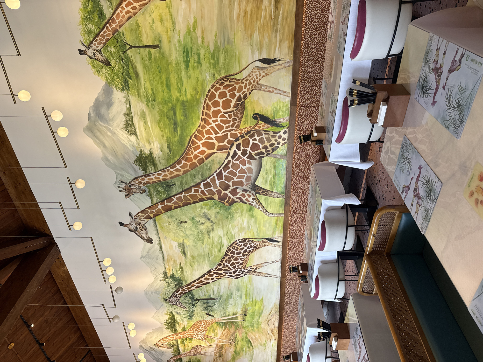 The image shows a restaurant dining area with a large mural of giraffes on the wall. The mural depicts several giraffes in a grassy landscape with mountains in the background. The restaurant has tables set with white tablecloths and chairs. Some tables have placemats with images of giraffes. The lighting fixtures are modern and minimalist. The overall decor suggests a safari theme.