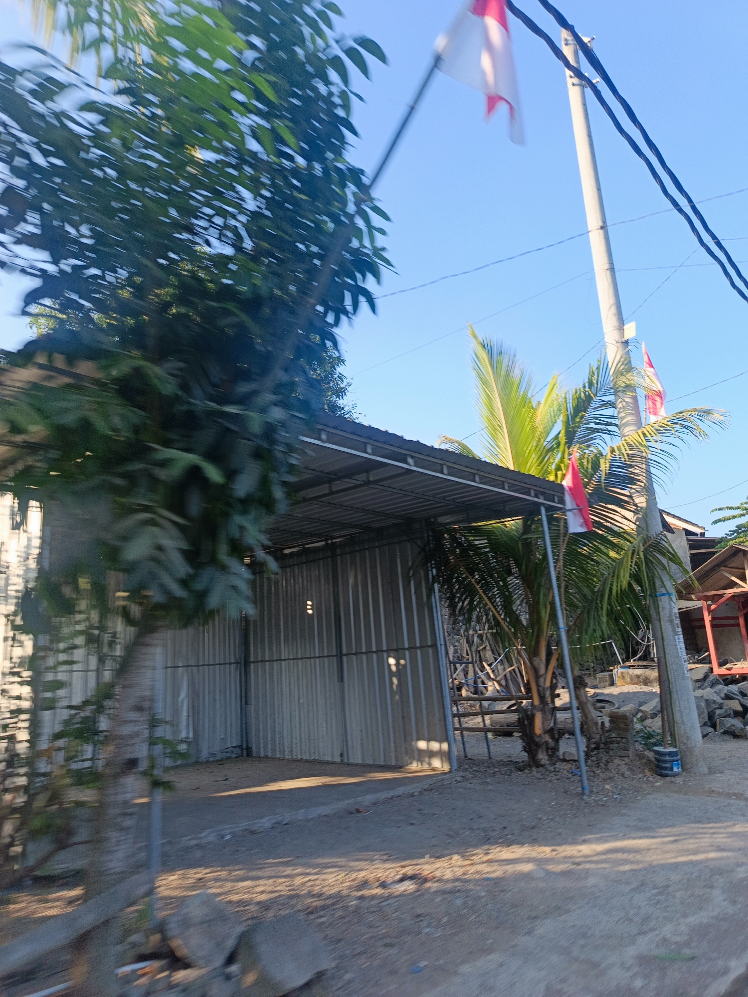 The image depicts an outdoor scene in Rembitan, Indonesia, under a clear blue sky. A prominent tall, light-colored utility pole stands on the right side of the frame, with black electrical wires running across the top. Two Indonesian flags, red and white, are visible; one is attached to the pole and waving, and another, smaller one is further down the pole and appears to be a smaller flag or decoration. In the foreground and mid-ground, there is a lot of foliage, with a large tree on the left side and palm trees and other greenery towards the right. The motion blur of the camera suggests the photo was taken from a moving vehicle.
A structure with a corrugated metal roof and walls is visible on the left side, appearing to be some form of covered open-air building or carport. The ground is a mixture of dirt, gravel, and some scattered rocks. Shadows are cast on the ground, indicating the time of day is likely morning or late afternoon. In the background, some smaller structures and piles of rocks are present, and vegetation is sparse. No people are clearly visible in the image.