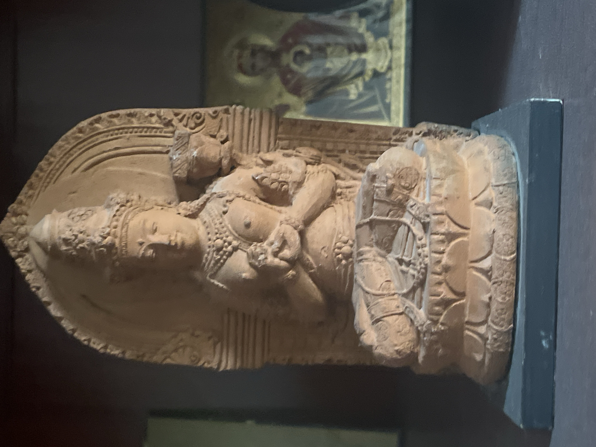 This is a close-up, slightly angled, vertical, eye-level, indoor shot of a small, terracotta statue of a seated deity. The deity is wearing an ornate crown, elaborate jewelry, and is in a meditative pose with hands clasped together at the chest. The statue is mounted on a lotus-shaped base, which itself rests on a dark, rectangular platform. Behind the statue, to the right, is a blurred icon of a religious figure, also appearing to be in a seated or standing posture. The background is dark and indistinct, suggesting it is a shelf or display in a dimly lit room. There is no visible indication of the city of Sochi, Russia, or any elements that would place the scene there.