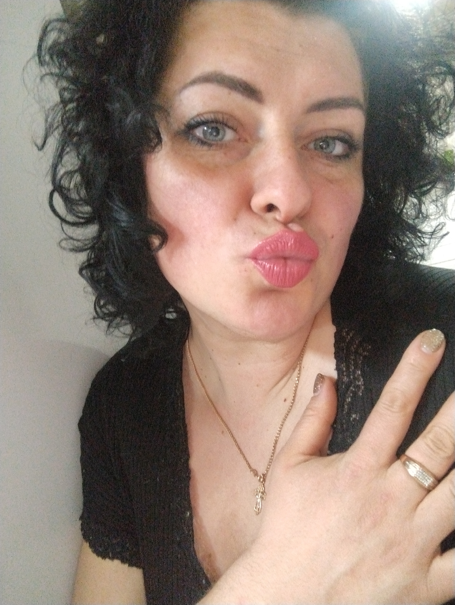 A woman is depicted in a close-up selfie, looking directly into the camera with a playful expression, her lips puckered into a kissing pose. She has dark, curly hair and striking blue-gray eyes, enhanced with eyeliner. She wears a black top that appears to have a knitted or lace texture. Around her neck is a golden chain with a small cross pendant. Her right hand is raised into the frame, revealing a gold ring with embedded stones on her ring finger, and her fingernails are painted with gold glitter polish. The background is a simple, light-colored indoor wall, suggesting a casual indoor setting. The image conveys a warm and affectionate mood. The location context for this media file is Grishanki, Russia.