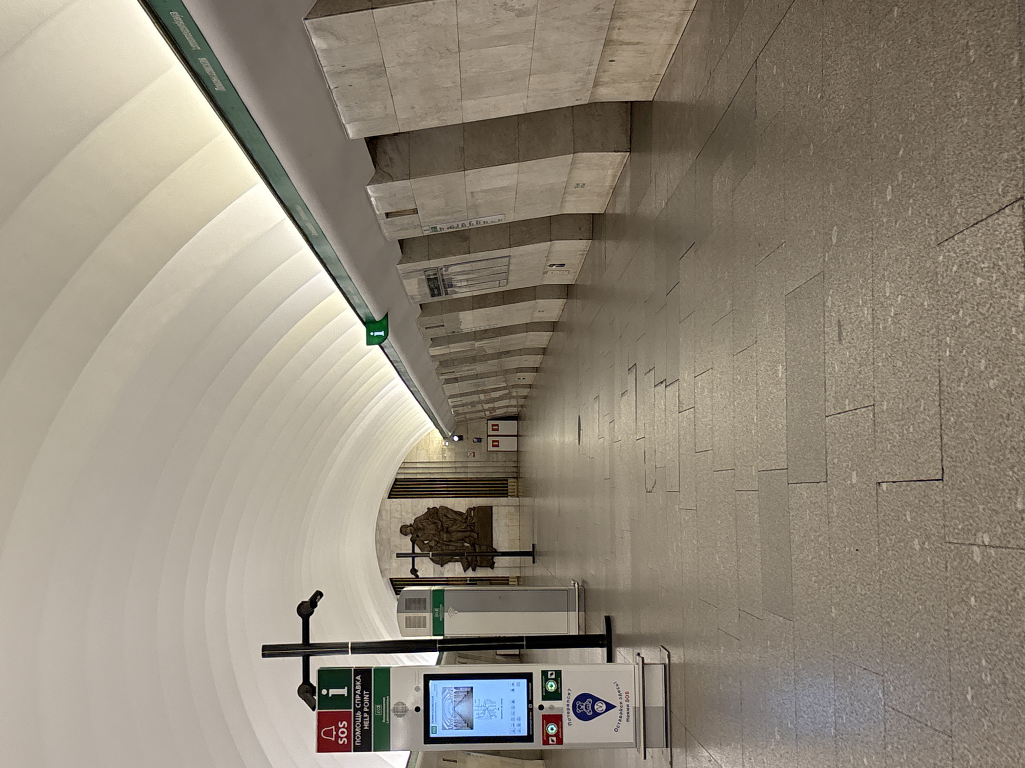 This image depicts the Lomonosovskaya metro station in Saint Petersburg, Russia. The scene captures an empty, long underground platform characterized by a distinct architectural style.
The station's ceiling is a prominent feature, consisting of a series of large, white, arched, and ribbed panels that run the entire length of the platform. Indirect lighting emanates from behind a continuous green linear fixture positioned along the base of the ceiling arch, which also displays the station name, "Lomonosovskaya," in both Cyrillic and Latin script.
The platform is supported by a row of imposing, trapezoidal pillars along the right side, constructed from light-colored, possibly marble or granite, stone cladding. These pillars angle slightly inwards as they ascend. The floor is tiled with light grey, speckled granite or terrazzo in a rectangular pattern, showing minor signs of wear and a few dark scuffs.
In the left foreground, a prominent white and black interactive "HELP POINT" kiosk stands. It features a digital screen displaying a metro map with "ЛОМОНОСОВСКАЯ" visible. Above the screen are a red "SOS" button (bell icon) and a green "i" (information) button. Below the screen, text reads "ПОМОЩЬ СПРАВКА HELP POINT," and further down, "Потерялся? Оставайся здесь! Нажми SOS" with a blue bear icon and a circular logo containing a "V". Behind this kiosk, a taller, narrower grey and white service terminal is visible. A black metal post with mounted components, possibly cameras or speakers, stands near the help point.
At the far end of the platform, a large bronze-toned sculptural composition is featured against a backdrop of vertical fluted wall panels. The sculpture appears to depict multiple figures, consistent with a theme of labor or science. Further down the platform on the right, two red service doors are visible. Small green "i" (information) signs are intermittently placed along the green ceiling strip. The station is devoid of people, presenting a quiet and monumental atmosphere.