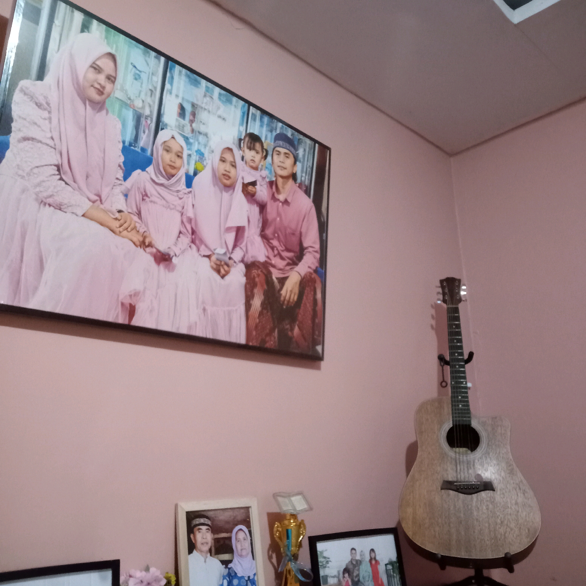 This image depicts an indoor scene, likely a living room, with a focus on a large framed photograph hanging on a pink wall. The photograph captures a family of six dressed in traditional Islamic attire, with the women and children wearing pink hijabs and dresses, and the man wearing a pink shirt and a patterned sarong. They are seated together, with the parents in the foreground and the children in the background. To the right of the photograph, an acoustic guitar is mounted on a stand against the same pink wall. In the foreground, on a surface below the large photograph, are several smaller framed pictures, a golden trophy with a blue ribbon, and what appears to be a small arrangement of flowers. The overall environment suggests a family's personal space, with decorations that indicate pride in family and possibly achievements. The presence of traditional attire in the photograph suggests a cultural context that is common in many parts of Indonesia, including Purwakarta.