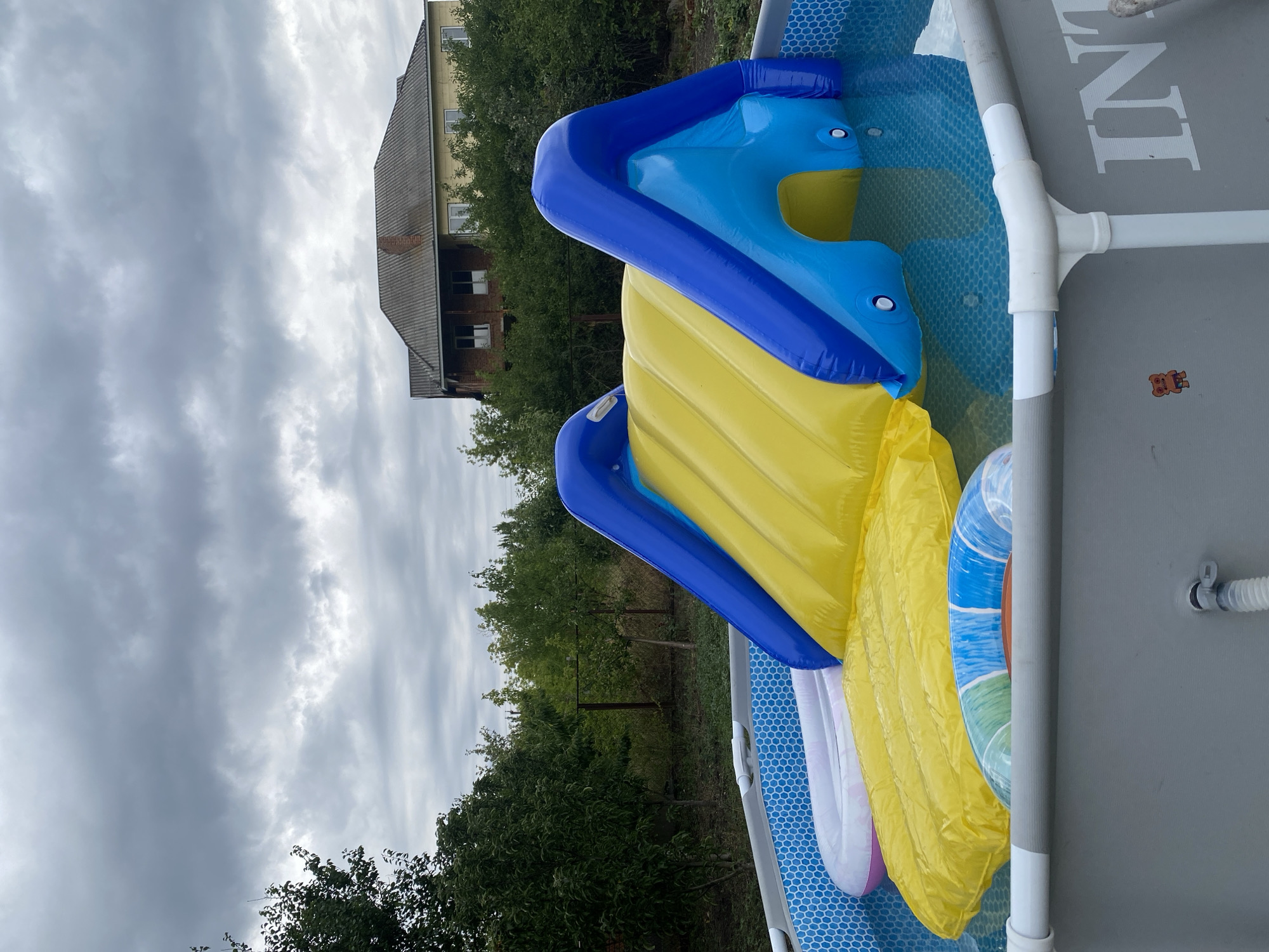 An inflatable water slide, predominantly yellow and blue, is set up in a rectangular above-ground swimming pool. The pool is filled with clear blue water and has a patterned liner. Floating in the pool are several inflatable items: a yellow raft and a circular inflatable ring with blue, green, and white stripes. The pool is surrounded by green foliage and trees. In the background, a large, two-story house with a grey roof and yellow siding is visible. The sky is overcast with heavy grey clouds. A small, cartoon-like sticker of a bear-like creature is affixed to the grey exterior wall of the pool on the right side. The location is Kropyvnytskyi, Ukraine.