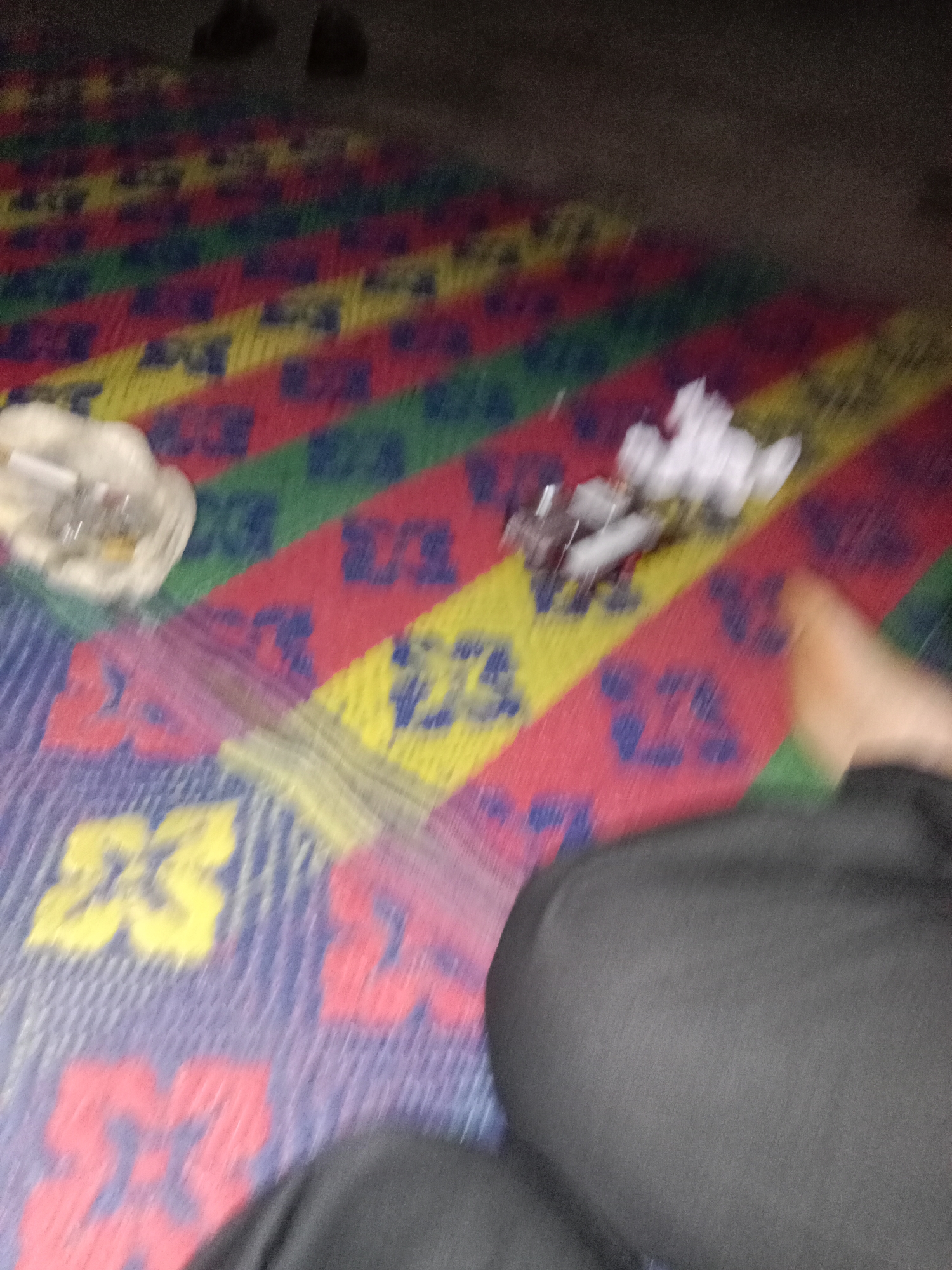 The image depicts a blurry, indoor scene with a focus on a colorful mat. The mat has diagonal stripes of red, yellow, blue, and green, with a repeating pattern of blue symbols on the red and yellow sections. In the upper left corner, a white object resembling an ashtray with cigarette butts and other debris is visible. To its right, a pile of white, crumpled paper or fabric is present. The lower portion of the image shows a person's leg and knee covered by dark clothing, and a bare foot. The background is dark and out of focus, with two dark, indistinct shapes that could be shoes or furniture. The overall lighting is dim, contributing to the blurriness of the image. No specific location in the city of Liwa, Indonesia can be determined from the visual information provided.