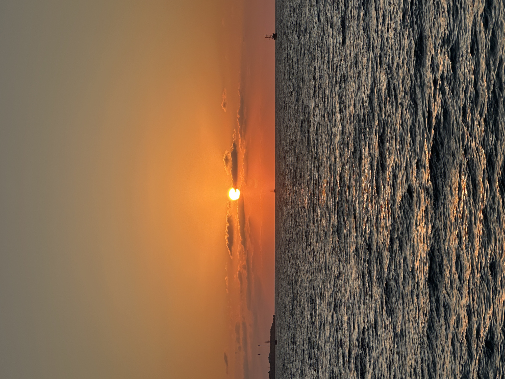 A seascape photograph depicts a sunset over the sea. The sky is a gradient of orange and yellow, with the sun partially obscured by clouds. The sea is choppy, with sunlight reflecting on the water. In the distance, there are a few sailboats and a landmass with buildings and flags, likely representing the coastline of Kyrenia, Northern Cyprus. The overall impression is one of tranquility and natural beauty.