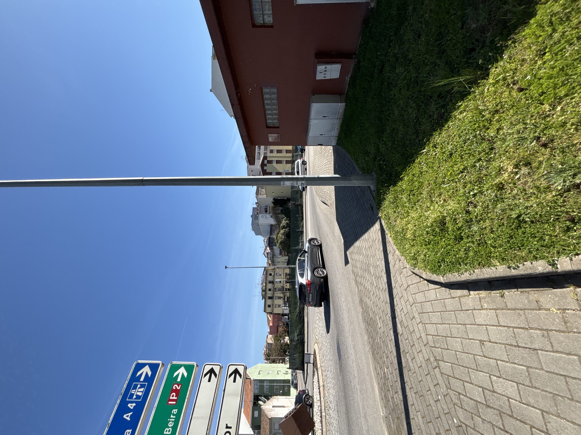 The image depicts a street scene in Mirandela, Portugal. A silver car is parked on the right side of the road, facing away from the camera. The street is paved, and a sidewalk runs along the right side, bordered by a grassy area. On the left side of the street, there are several road signs, including one for "A4" and another for "Beira IP2." A tall, silver lamppost stands to the left of the road signs. In the background, there are buildings of varying heights and colors. The sky is clear and blue. A red building is on the right side behind the grass.