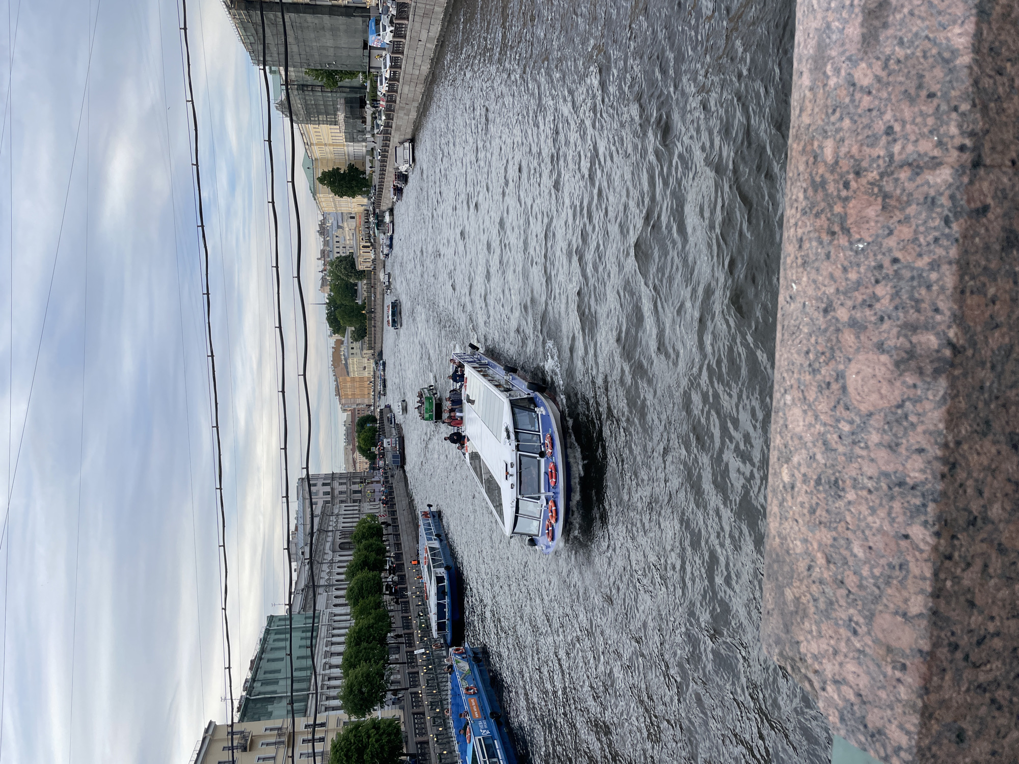The image presents an urban river or canal scene, captured from an elevated perspective, likely a bridge. The scene is oriented vertically within the frame, with the sky on the left, the river and buildings in the middle, and a granite structure on the right.
**Subjects:**
The central focus is a busy river, dark and rippled, where several passenger boats are actively navigating. The most prominent boat, positioned slightly right of the center, is white and blue with a glass-enclosed upper deck and open seating areas; it carries numerous people. Orange life rings are visible on its side. Other smaller, darker blue boats are also seen further along the waterway. Lining the far bank of the river is a collection of historic multi-story buildings, varying in architectural style and color (creams, greys, and yellows), interspersed with green trees. The left portion of the image is dominated by a pale sky with soft white and grey clouds. Several dark electrical or communication wires stretch horizontally across the sky. On the far right, a large section of a brown and black speckled granite structure, likely a bridge railing or pillar, frames the scene.
**Activity or event taking place:**
The boats are engaged in active transit on the water, indicating a sightseeing tour or transportation service. Passengers are visible on the decks, suggesting a leisure activity.
**Notable details:**
The time of day appears to be daytime, with a soft, diffused light suggesting either an overcast sky or early morning/late afternoon conditions. The weather is partly cloudy to overcast. The architectural style of the buildings, the presence of numerous canals, and the type of tour boats are consistent with a major European city like Saint Petersburg, Russia, the specified location. The overall atmosphere is one of calm urban activity and tourism.
**Visible text:**
On the side of the prominent white and blue boat, the Russian word "ЛЕТО" (LETO), which translates to "SUMMER," is clearly visible in white lettering.