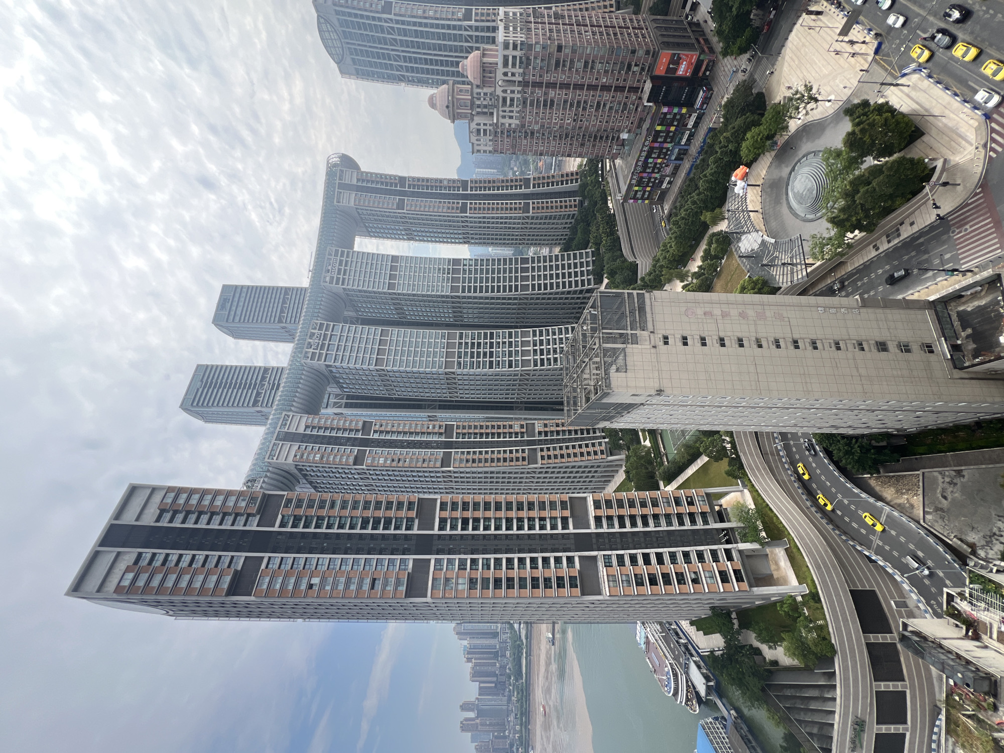 This image shows a panoramic view of the Yuzhong District in Chongqing, China, featuring a collection of towering skyscrapers and a distinctive connecting structure. The "Crystal" building, a prominent architectural element, consists of multiple vertical towers linked by a horizontal glass-enclosed structure. The foreground is dominated by a curving elevated road with several yellow vehicles, including taxis, and a silver car. Adjacent to the road is a large, multi-story building with a grey facade and a complex street-level commercial area featuring colorful signage. Further into the scene, the Yangtze River is visible with a large cruise ship docked along its bank. In the background, a cityscape of numerous high-rise buildings stretches across the horizon under an overcast sky. The overall environment is urban, characterized by dense construction and significant infrastructure.