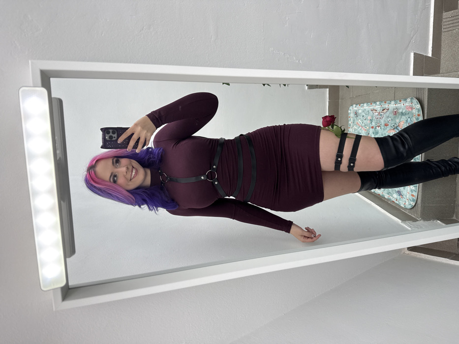 A female individual is captured in a mirror selfie inside what appears to be an indoor setting in the City of Zagreb, Croatia. The subject, a woman, stands facing the mirror, holding a smartphone with a black cat-eared case in her left hand to take the photograph. She has long, wavy hair with pink roots transitioning to purple ends. Her facial expression is a smile, with her head slightly tilted. She is dressed in a form-fitting, long-sleeved, high-neck maroon mini-dress. Over the dress, she wears a black leather harness with silver rings and straps around her torso. A matching black leather double-strap garter is secured on her right thigh, with a single red rose tucked into it. She is wearing black thigh-high boots. Above the mirror, a white light fixture with multiple illuminated bulbs is visible. The immediate environment includes plain white walls and a light grey floor, where a light blue mat with a subtle pattern is situated.