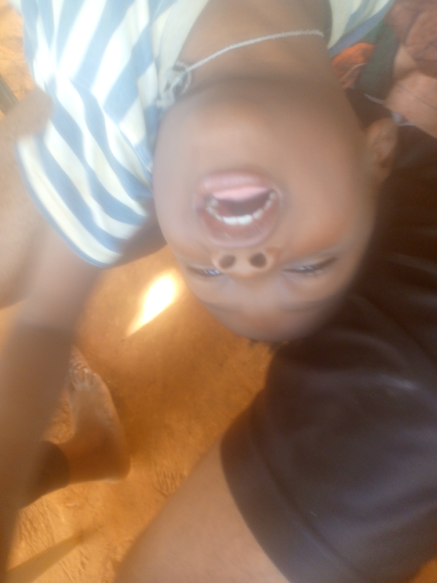A child, with dark skin, is turned upside down. They are wearing a blue and white striped shirt and a silver necklace. Their mouth is open, revealing their teeth and tongue. The child is lying on a brown surface. Part of a black garment is also visible. The location is unknown.