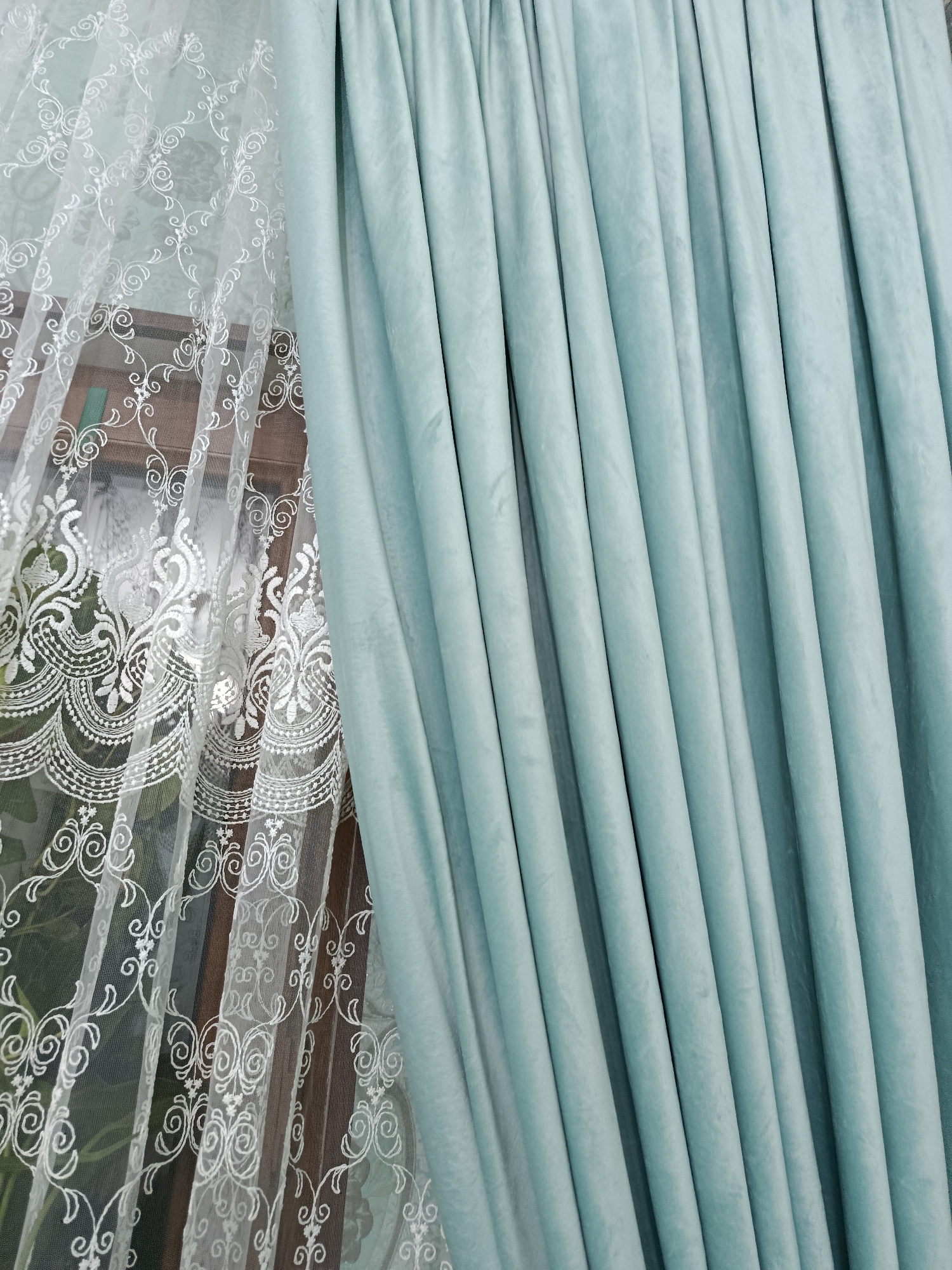 A close-up shot shows a pair of curtains with one side being a sheer white lace with an ornate floral pattern and the other side being a solid teal velvet curtain. The teal curtain exhibits a rich texture and is neatly pleated, suggesting it is draped. The lace curtain is semi-transparent, allowing a blurred view of what appears to be window panes and possibly greenery behind it. The composition is focused on the textures and colors of the drapery, with no people or specific landmarks visible to indicate a location in Tashkent, Uzbekistan.
