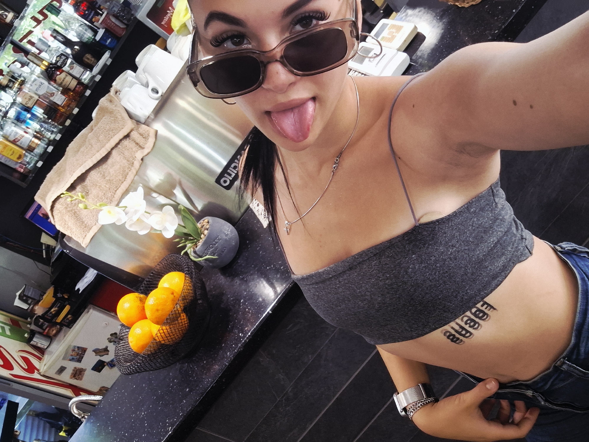 An indoor, high-angle selfie captures a young woman in what appears to be a cafe or bar setting in Mezdra, Bulgaria. She is positioned slightly to the right, facing the camera with a playful expression, sticking her tongue out. Her dark hair is pulled back, and she wears rectangular, translucent brown sunglasses that obscure her eyes but reveal long, dark eyelashes.
She is dressed casually in a gray strapless bandeau top and blue jeans, which are unbuttoned and partially pulled down on her left side. A silver chain necklace with a small cross pendant hangs around her neck. On her left wrist, she wears a silver-toned metal watch and a delicate bracelet. A prominent stylized black tattoo reading "BUBBLE" or a similar word is visible on her right side, just below her ribcage.
The background features a well-stocked bar or countertop area. To her left, a black mesh basket holds several bright oranges. Beside it, a small gray potted plant with white artificial orchid flowers adds a decorative touch. A beige folded towel rests on a stainless steel surface, where white ceramic coffee cups are stacked, suggesting the presence of a coffee machine. Behind these, shelves display numerous bottles of various liquors and water. Part of a white tag with the word "OLIDIO" is visible hanging near the woman's head. The countertops are dark, possibly black granite or speckled laminate. The overall ambiance is relaxed and casual.