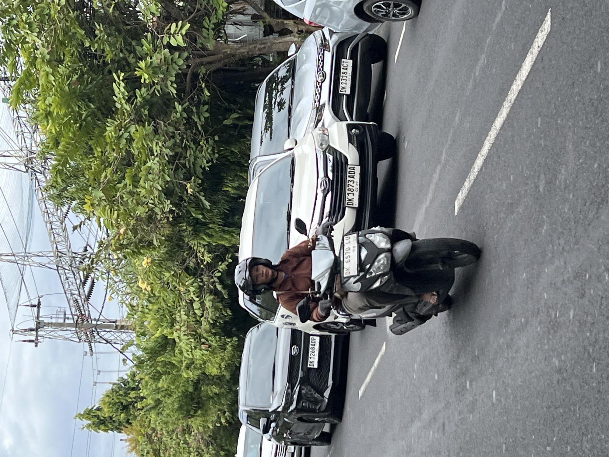 In Denpasar, Indonesia, a person wearing a helmet and a brown jacket rides a scooter on a road. The scooter has a license plate that reads "DK 6570 LU." Several cars are also visible on the road, including a black car with a license plate "DK 1268 ADP," a white car with a license plate "DK 1873 ADA," and a silver car with a license plate "DK 1318 ACT." In the background, there are lush green trees and power lines overhead. The sky is overcast.