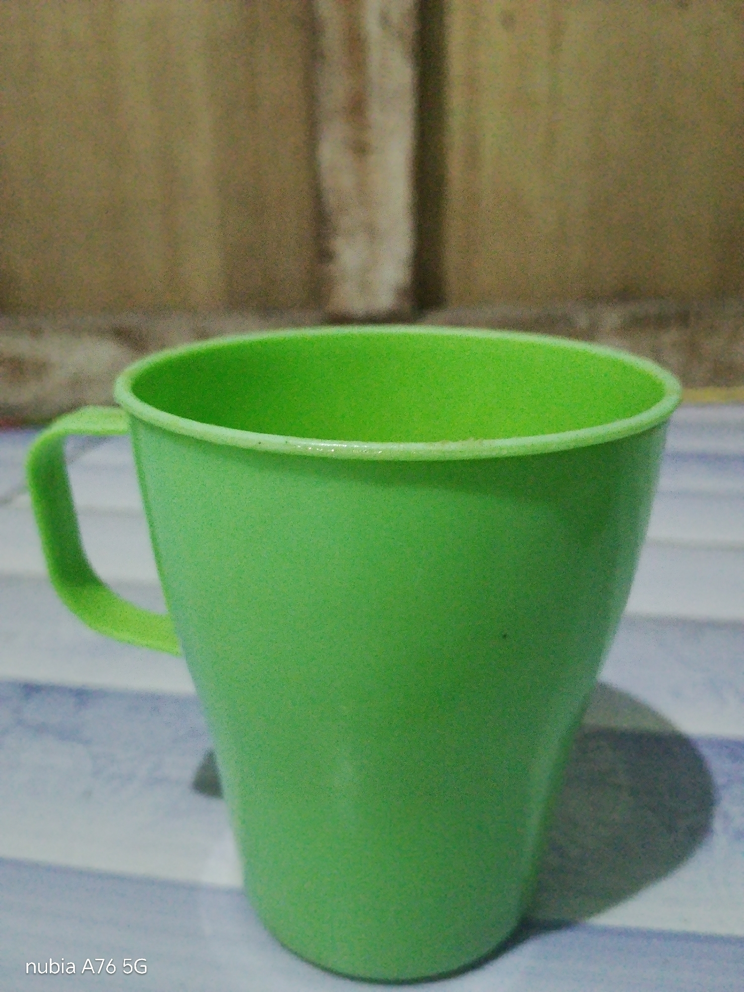 A bright green plastic mug with a handle is positioned on a surface with a striped pattern. The mug is the primary subject, taking up most of the frame. The background is a textured, light brown wall. The lighting suggests an indoor setting, possibly a home or a casual establishment, and the overall scene is likely located in Binuang, Indonesia, as indicated by the "nubia A76 5G" watermark, which is a smartphone model often associated with region-specific branding. There are no people or visible actions occurring in the image. The mug casts a soft shadow to its right.