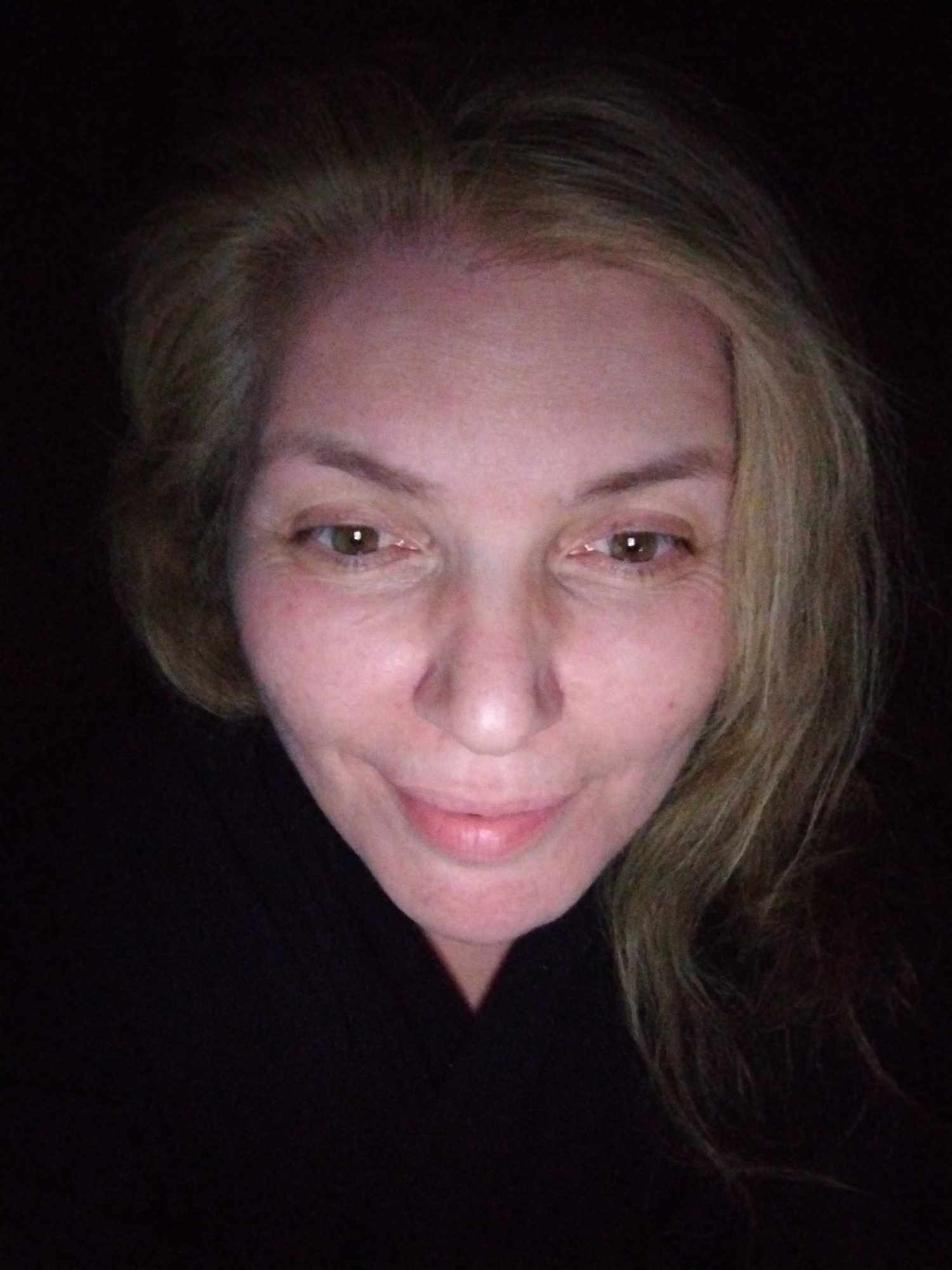 A close-up, dimly lit photograph shows a woman with fair skin and light-colored hair. She has brown eyes and a slight smile. She is wearing a dark garment. The background is dark and indistinct. There is no information about a city or the United Arab Emirates.