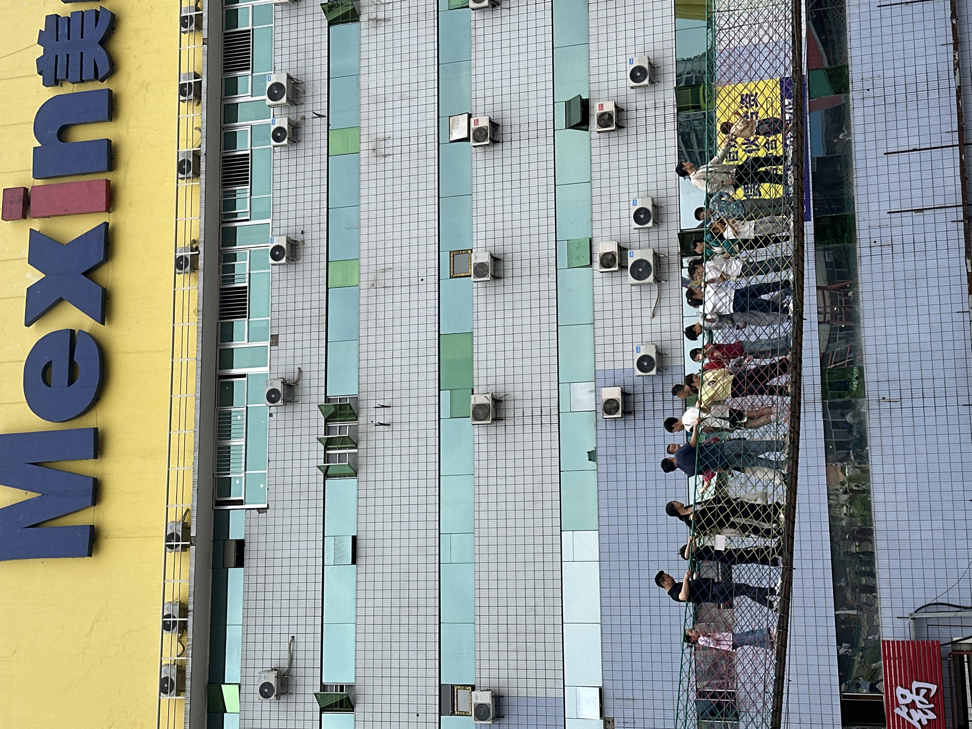 An eye-level shot captures a vibrant urban scene in Nan'an District, China, dominated by the side of a tall building. The building features a large yellow section on the left with prominent blue and red dimensional letters spelling "UNIX 美" (UNIX and the Chinese character 'Mei', meaning 'beauty' or 'beautiful'). To the right, the building's facade is covered in a light gray grid-patterned tiling, interspersed with vertical strips of light blue and occasional light green panels. Numerous white outdoor air conditioning units are mounted irregularly across this tiled section.
On the far right, a long, narrow platform or scaffolding ascends the building, enclosed by a beige and yellow mesh fence. Several people, mostly adults, are standing on this platform, appearing to observe or wait. Some individuals are seen holding onto the mesh. Below the platform, a reflective surface, likely a large window or panel on a lower part of the building, mirrors reflections of people, a yellow sign with Chinese characters (including '美'), and a red sign at the bottom right corner displaying another Chinese character, possibly '品'. The lighting suggests it is daytime, likely overcast, with soft shadows. The overall impression is that of a bustling urban environment.