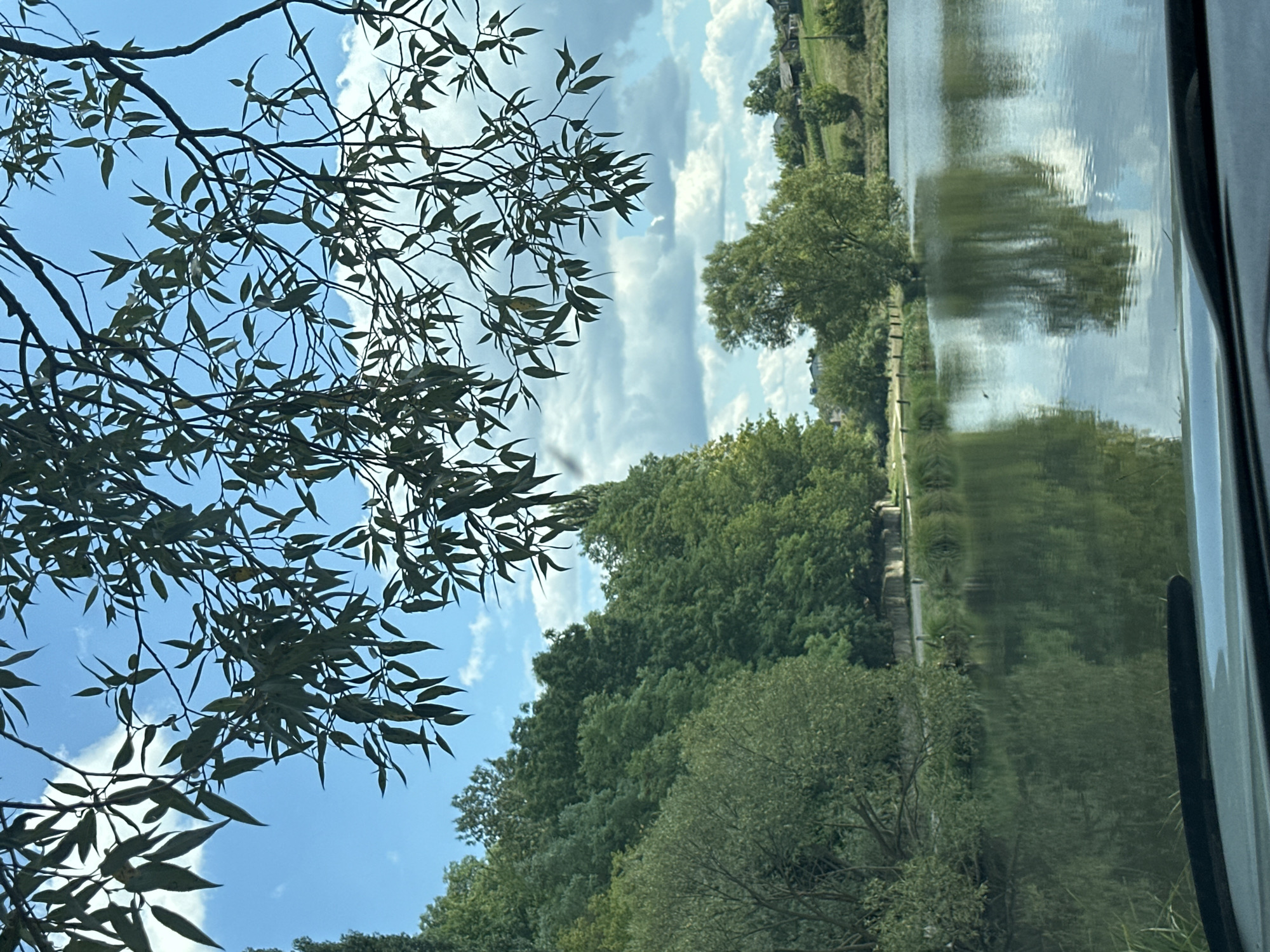 This is a photograph taken from within a vehicle, looking out at a serene body of water surrounded by lush green trees and a blue sky with scattered clouds. The perspective is from a low angle, showing the dashboard of the car in the foreground. The water reflects the trees and sky, creating a mirror-like effect. In the distance, a gentle slope leads up to several houses nestled amongst more trees. This scene is likely located in the vicinity of the village of Makiv, Ukraine, given the natural, rural setting. There are no people or animals visible in the image, and no discernible actions or interactions taking place. The environment is peaceful and natural, dominated by greenery and water.